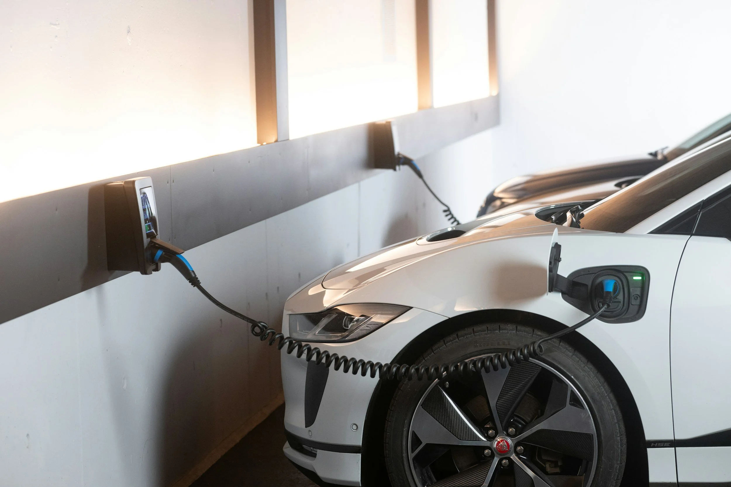 A white electric Jaguar I-PACE parked in a garage, connected to a charging station on the wall.