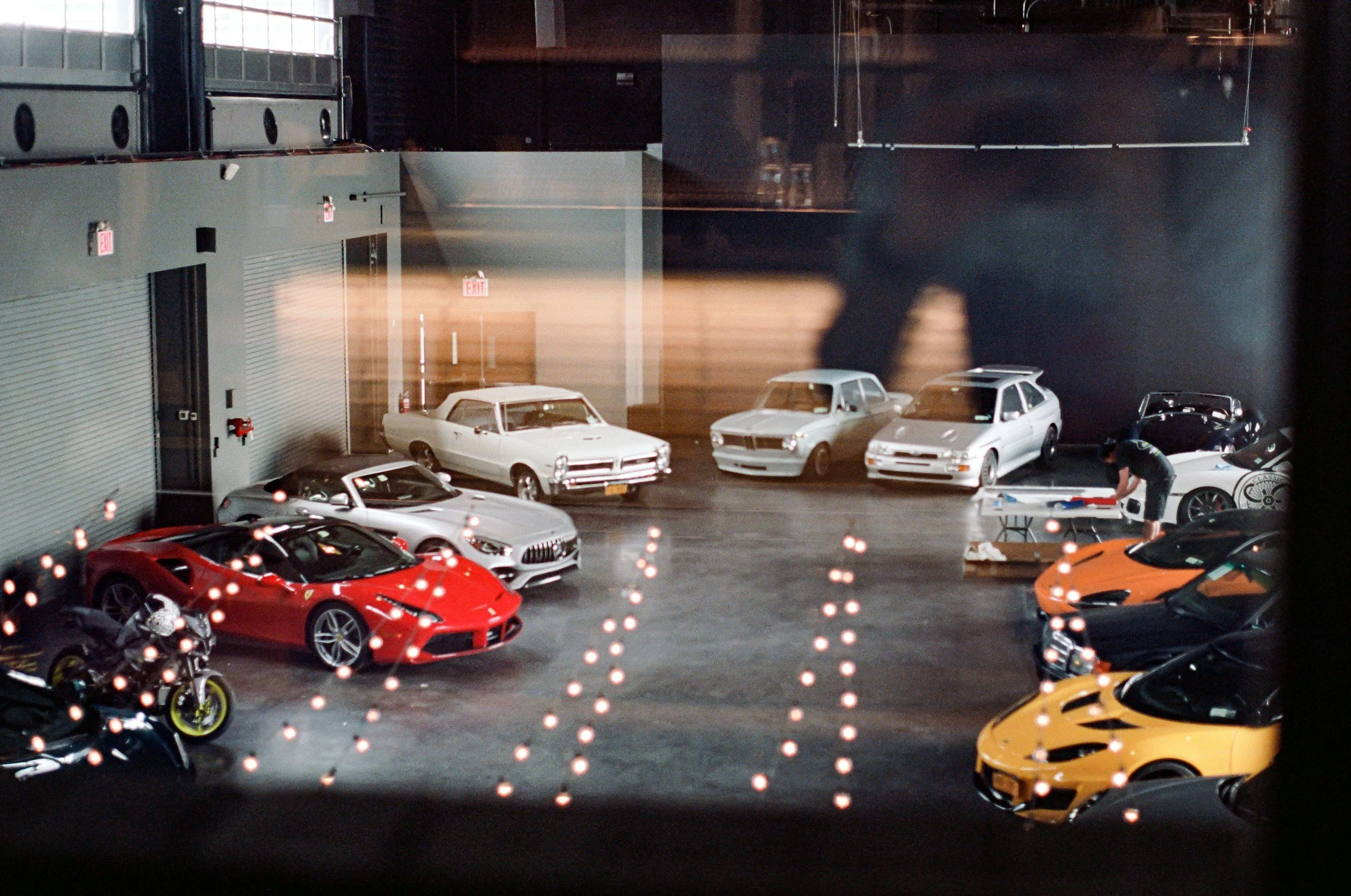 View of a car dealership with various sports cars, including a red Ferrari and an orange Lamborghini, seen through a window with reflections of lights.