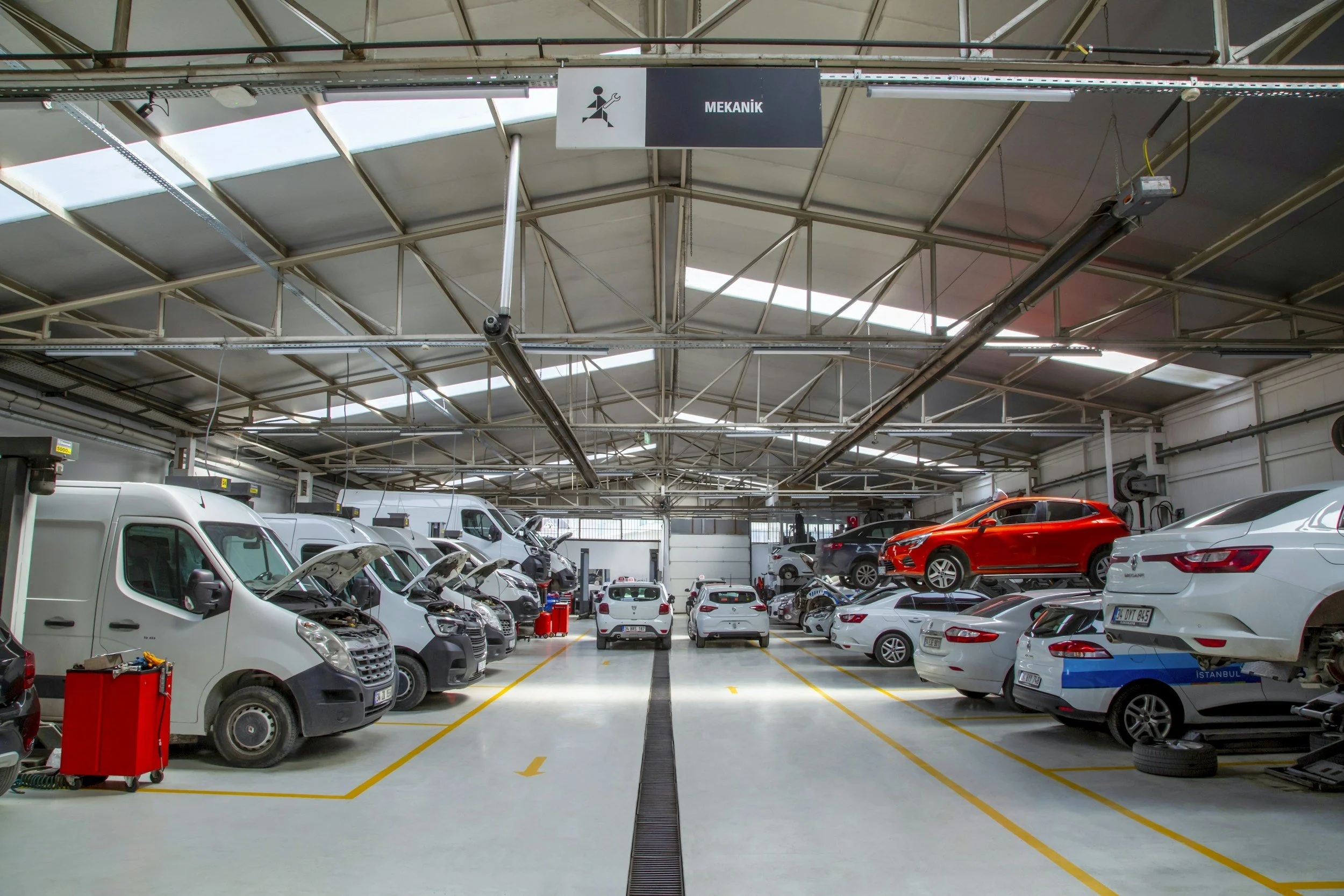 A well-lit indoor parking garage with cars parked on both sides, including a white van in the foreground and a red car on the right. Signs indicating mechanics are visible, and the ceiling has skylights and metal framework.