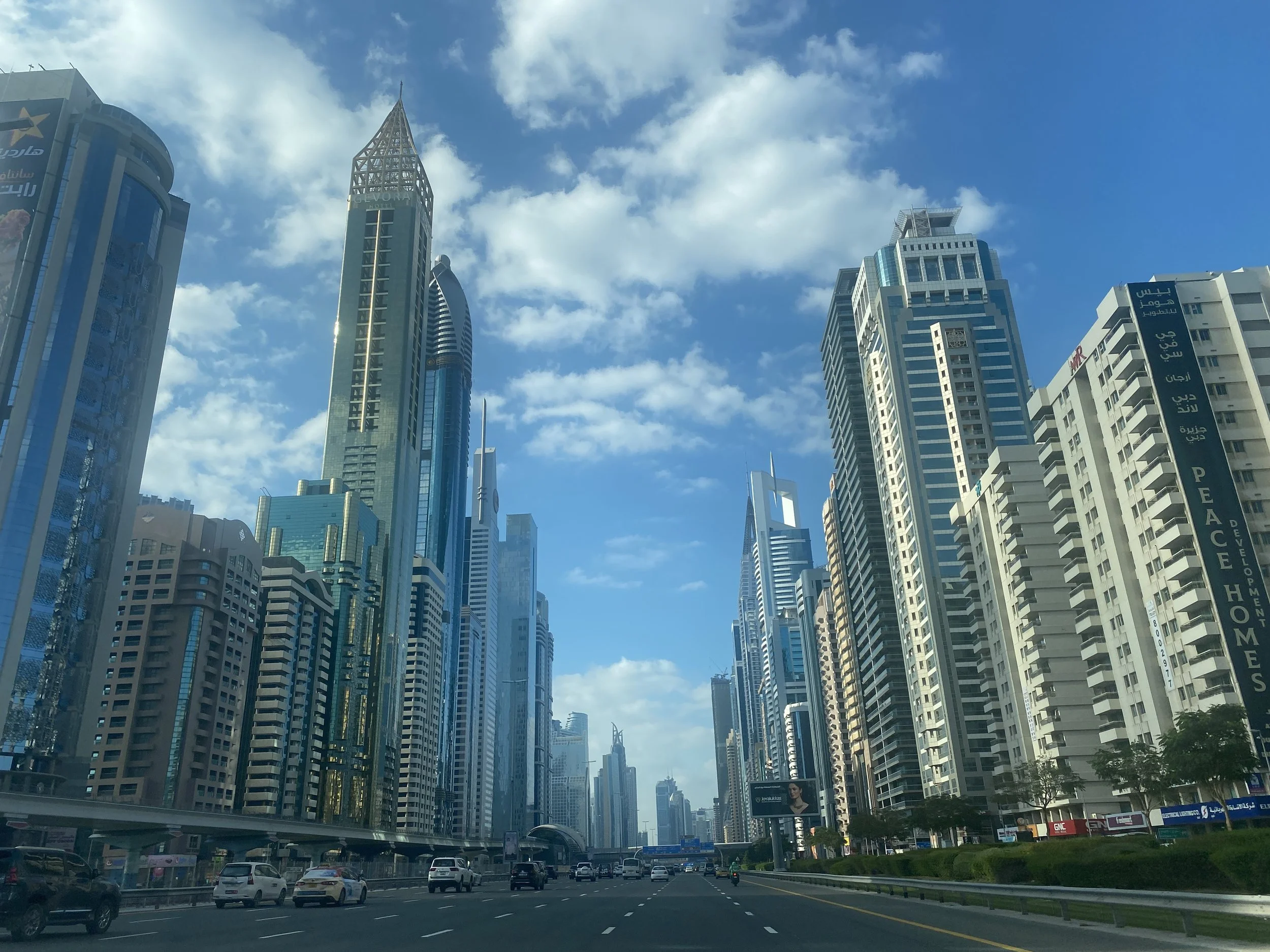 Dubai cityscape with tall modern skyscrapers, busy street with vehicles, billboards, and blue sky with scattered clouds.