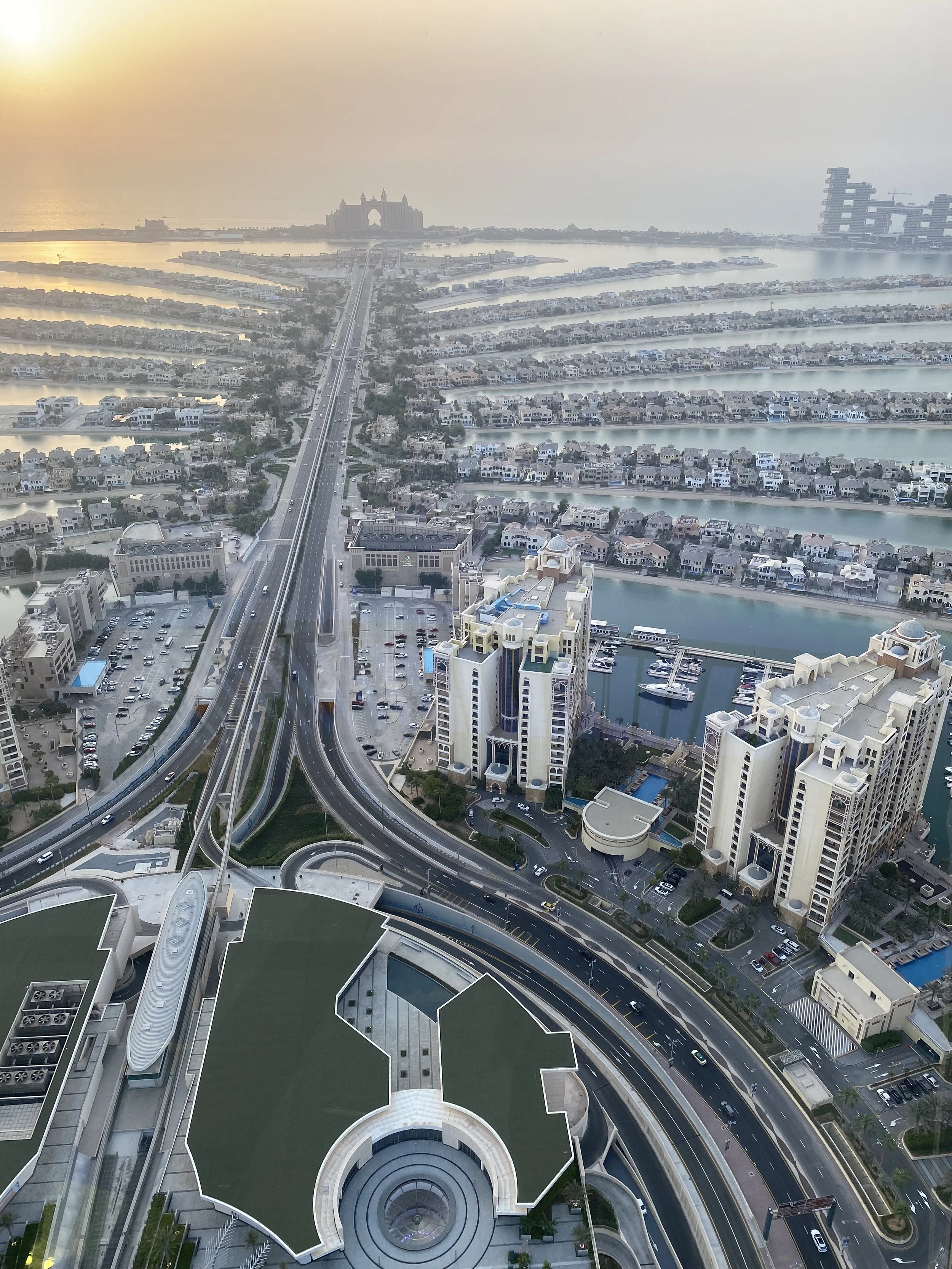 High-angle view of the Palm island showing a large building with a green roof, surrounded by roads, smaller buildings, and water bodies. The background includes the Atlantis The Palm hotel and the Atlantis The Royal hotel in Dubai.