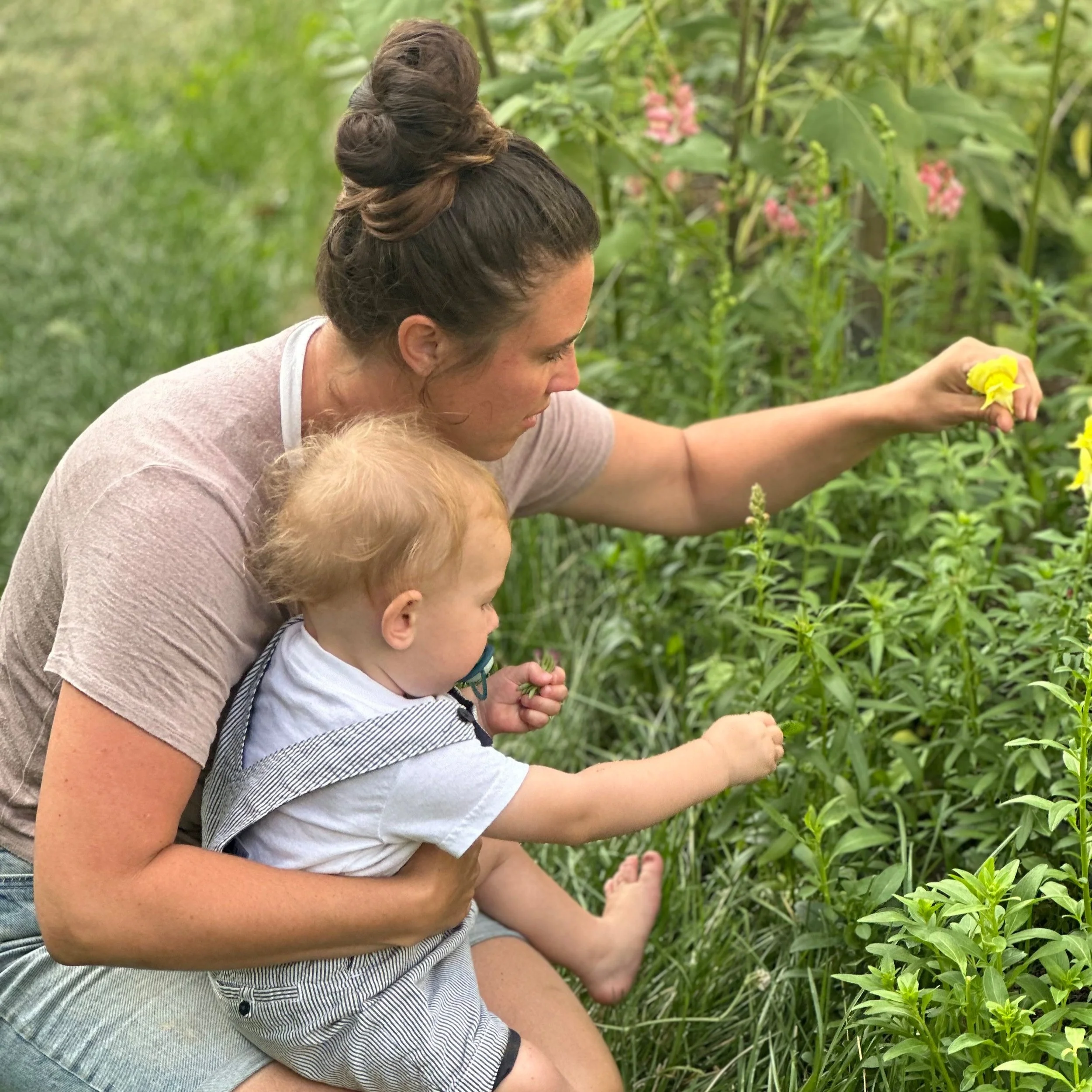 A woman with dark hair tied up in a bun holding a young child while they pick yellow flowers from a garden or field with green plants and pink flowers in the background.