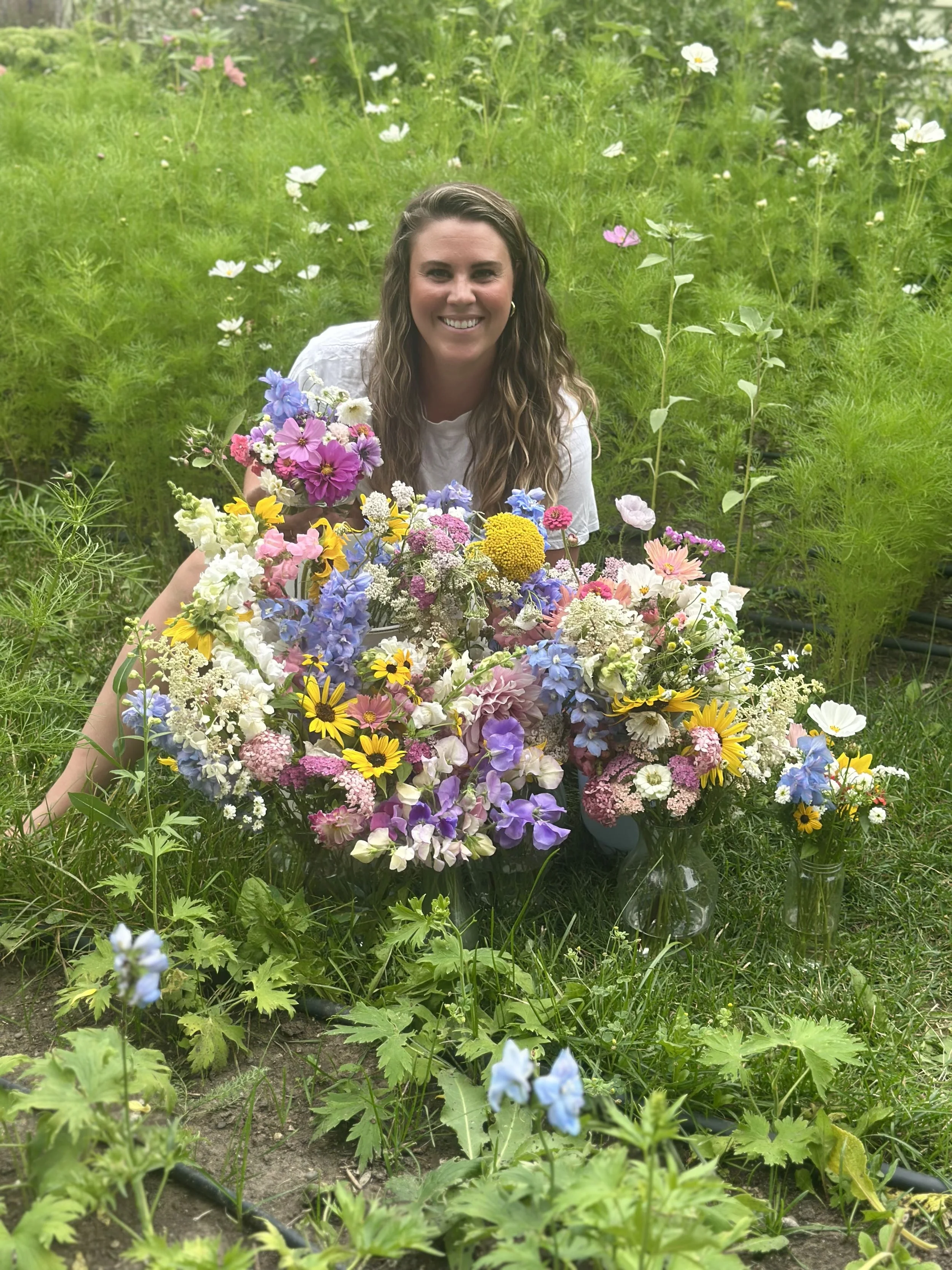 A woman with wavy brown hair smiling while surrounded by colorful flowers in a garden.