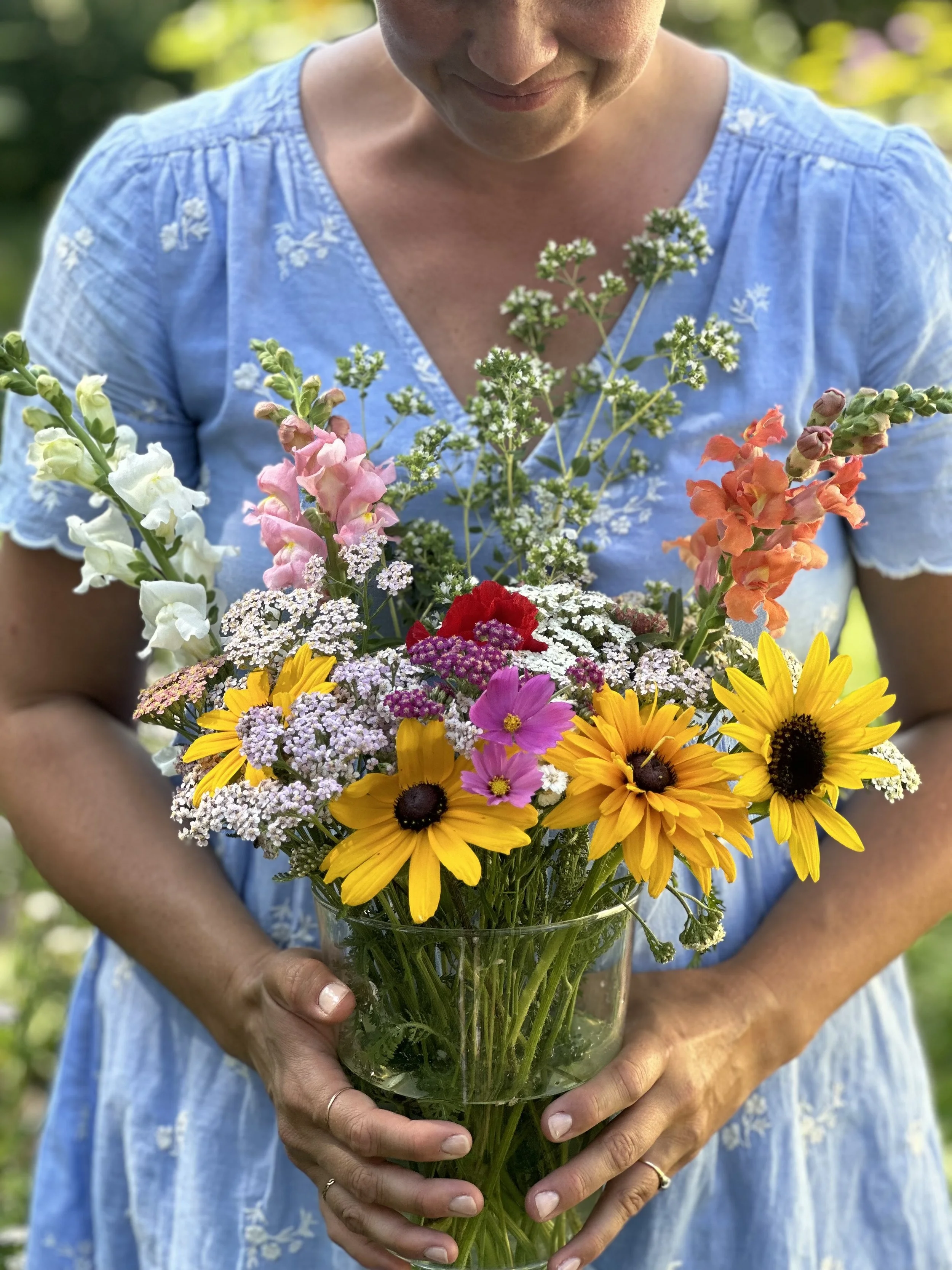 Person in a blue dress holding a vase of colorful fresh flowers, including sunflowers, pink cosmos, white snapdragons, and other wildflowers, outdoors with greenery in the background.