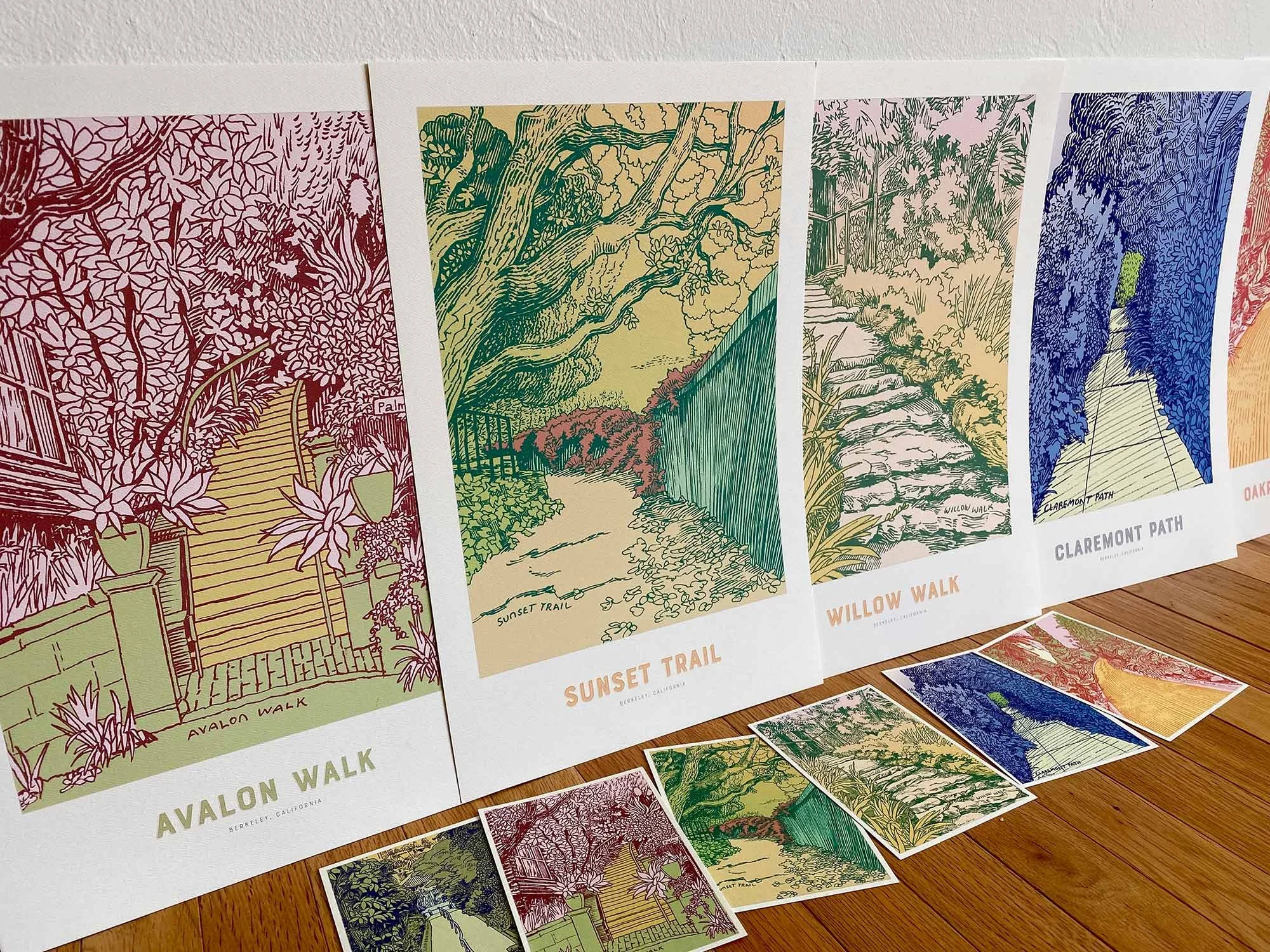 Four posters in a row of path artwork including Avalon Walk, Sunset Trail, Willow Walk and Claremont Path, and their corresponding postcards are spread out in front of them.