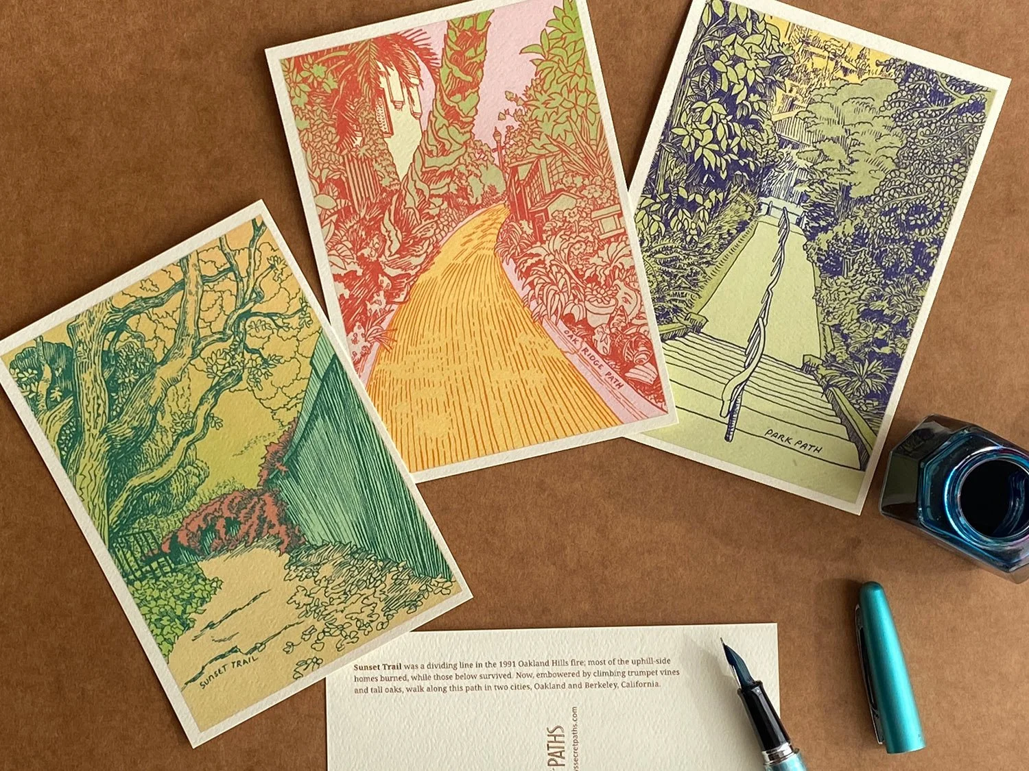 Three illustrated path postcards with a fountain pen and blue ink on a writing desk. Paths include Sunset Trail, Oakridge Path, and Park Path in Berkeley, California.