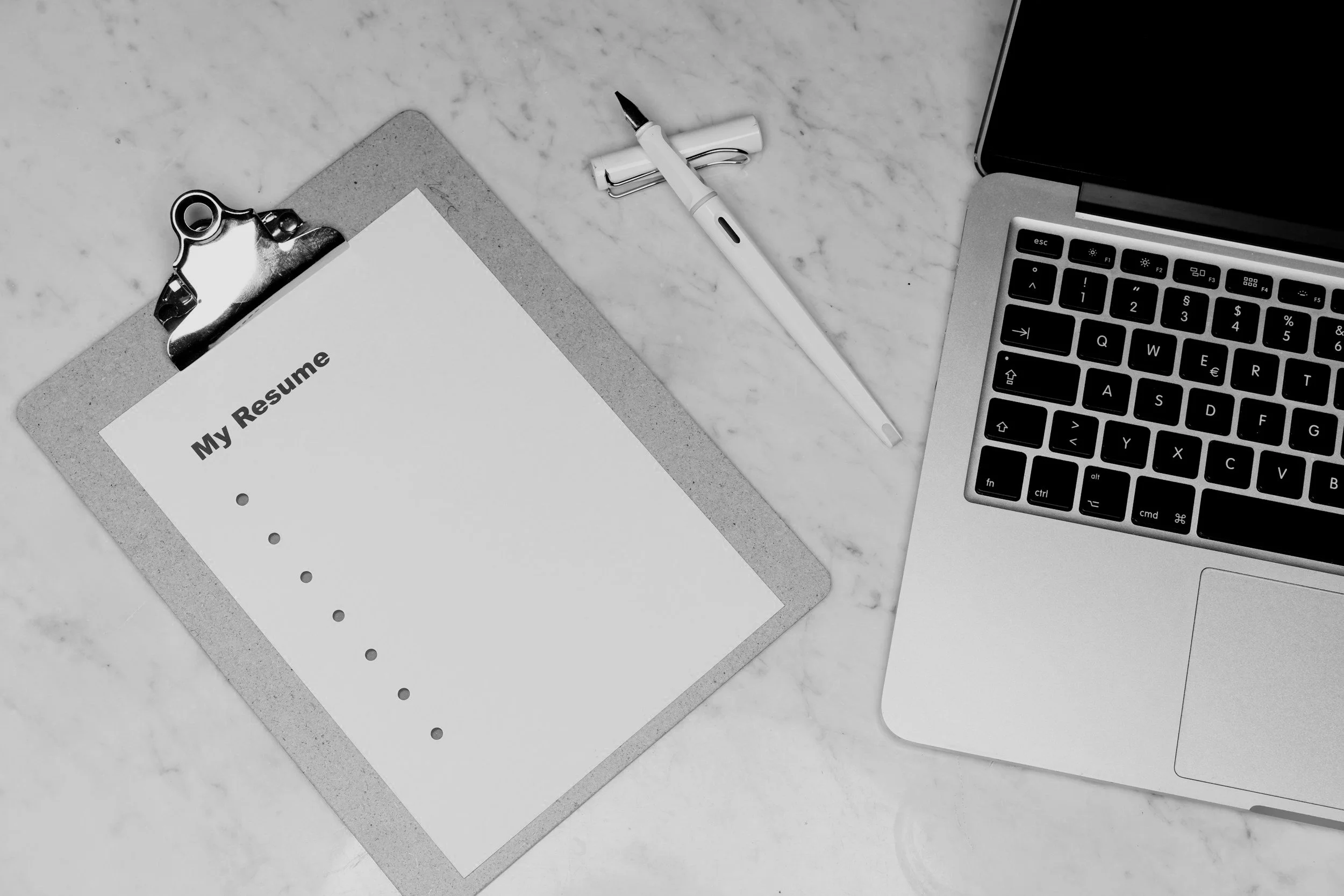 A workspace with a clipboard holding a resume, a white pen, and a laptop on a white marble surface.