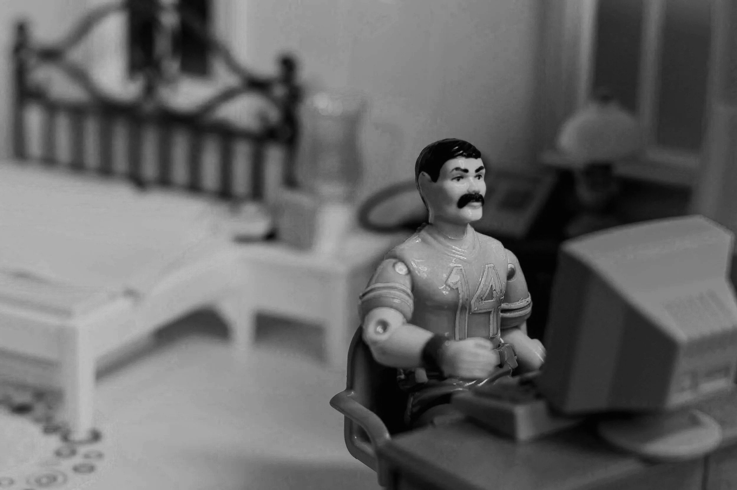 A small doll sits at a tiny desk, staring intently at a blank, dummy computer screen while searching for a new job.
