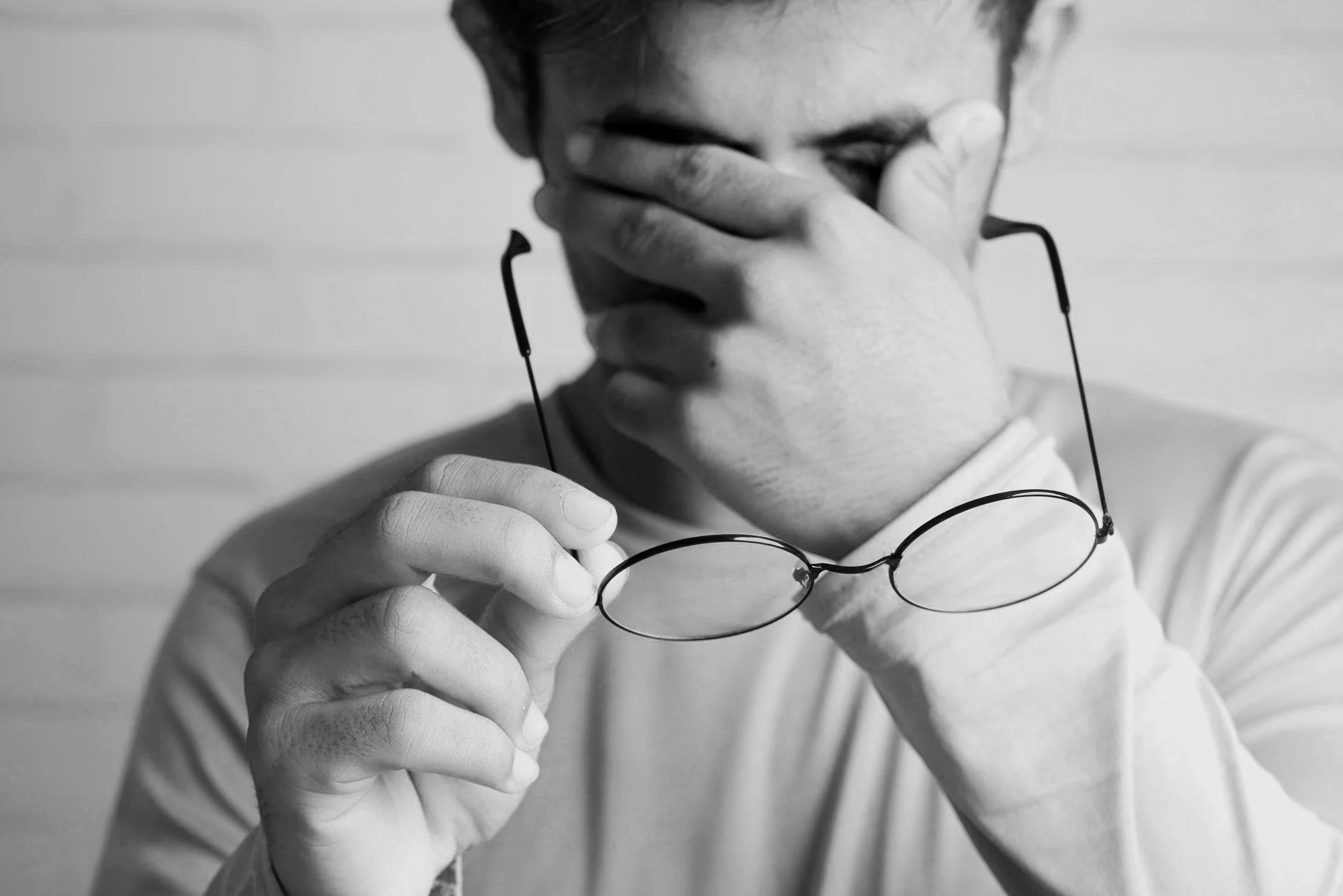 A person with glasses covers their face with one hand while holding their glasses in the other hand, in black and white.