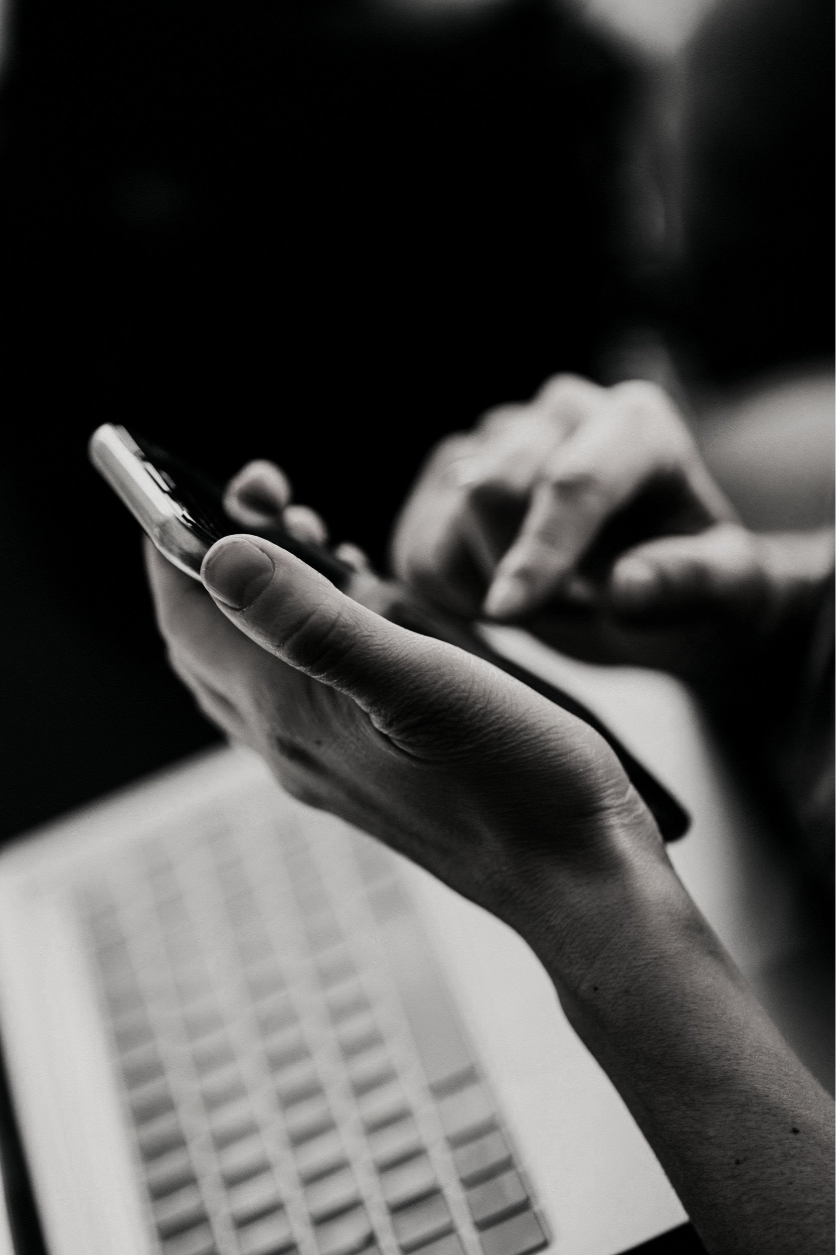 Close-up of person's hand holding a smartphone, with a laptop visible in the background, black and white photo.