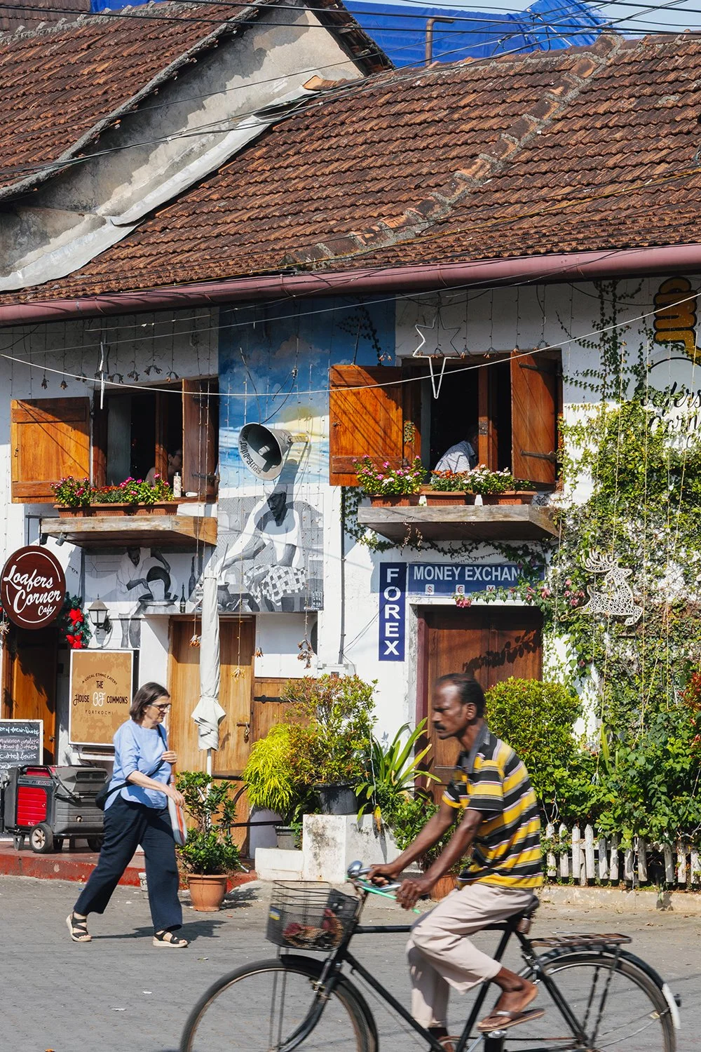 A street scene featuring a building with a mural, potted plants, and open windows with wooden shutters. A person is riding a bicycle, and a woman is walking nearby.