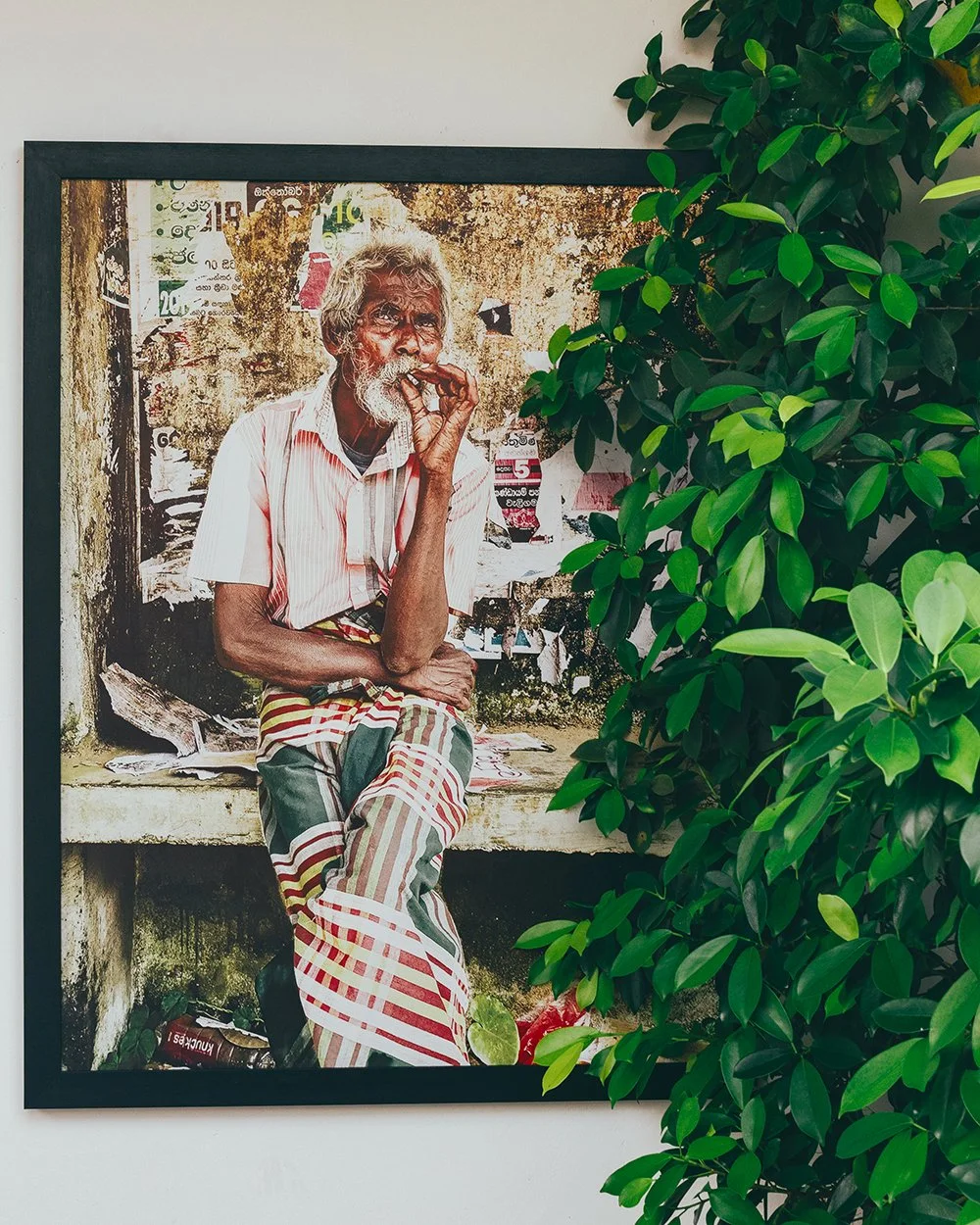 A framed photograph of an elderly man with gray hair and a beard, sitting against a weathered wall covered with torn posters. The man is wearing a light-colored, striped shirt and a traditional patterned sarong, and appears to be smoking a cigarette.