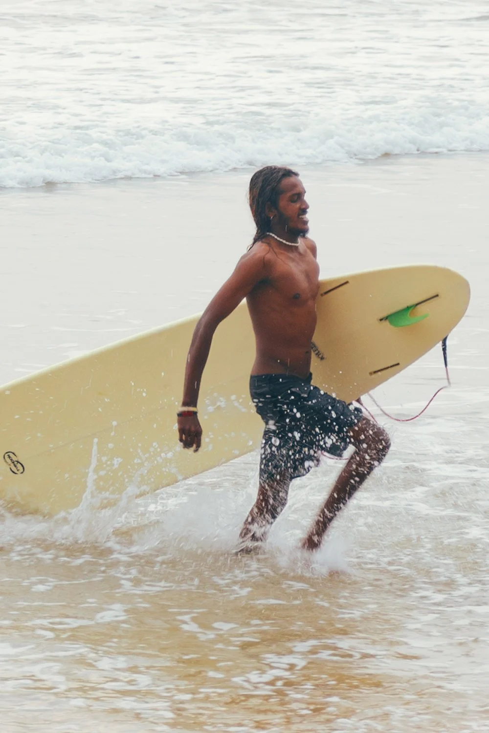 A man with long hair, wearing black board shorts, carrying a yellow surfboard while walking in shallow ocean water.