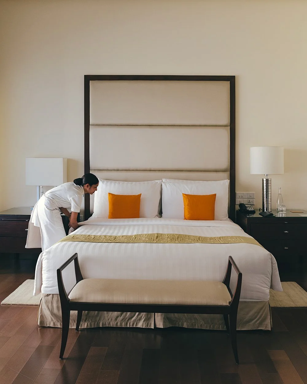 A hotel room with a large bed, two orange pillows, a padded headboard, a bench at the foot of the bed, and a woman in a white uniform tidying the bed. There are nightstands on both sides of the bed with lamps, a telephone, and a glass bottle with a g