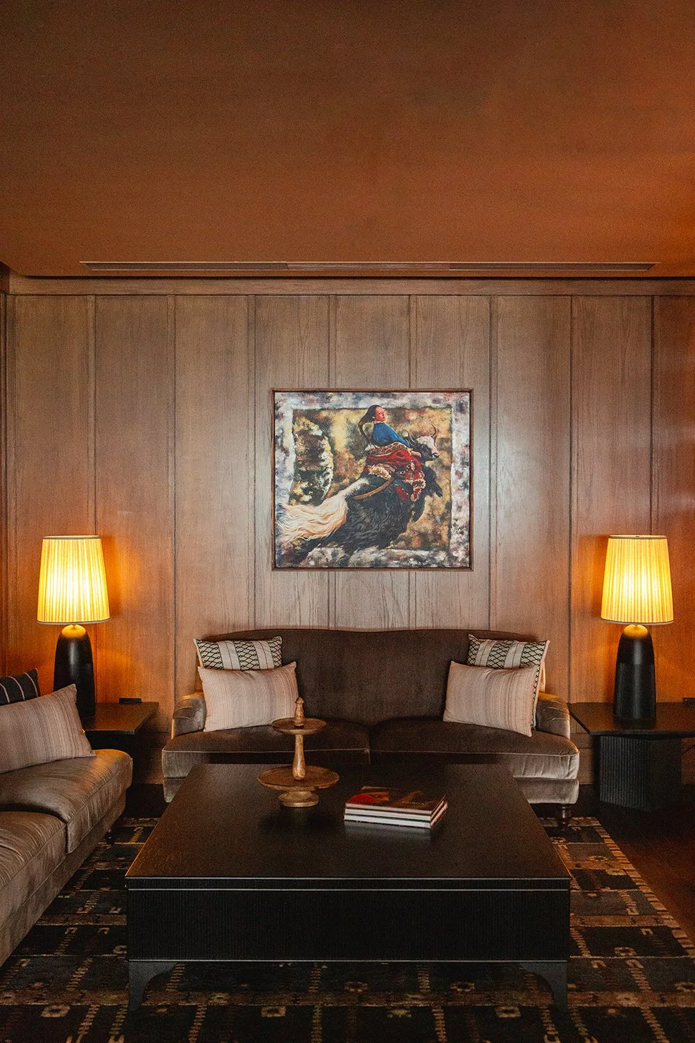 Living room with wood-paneled wall, two lamps on side tables, a dark sofa with pillows, a coffee table, and a painting of a woman riding a horse on the wall.