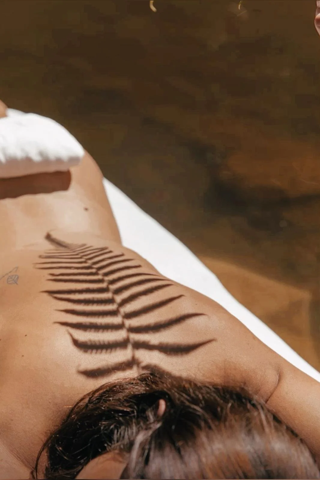A person's arm with a shadow of a fern leaf cast on it, lying on a white surface with a wooden background.