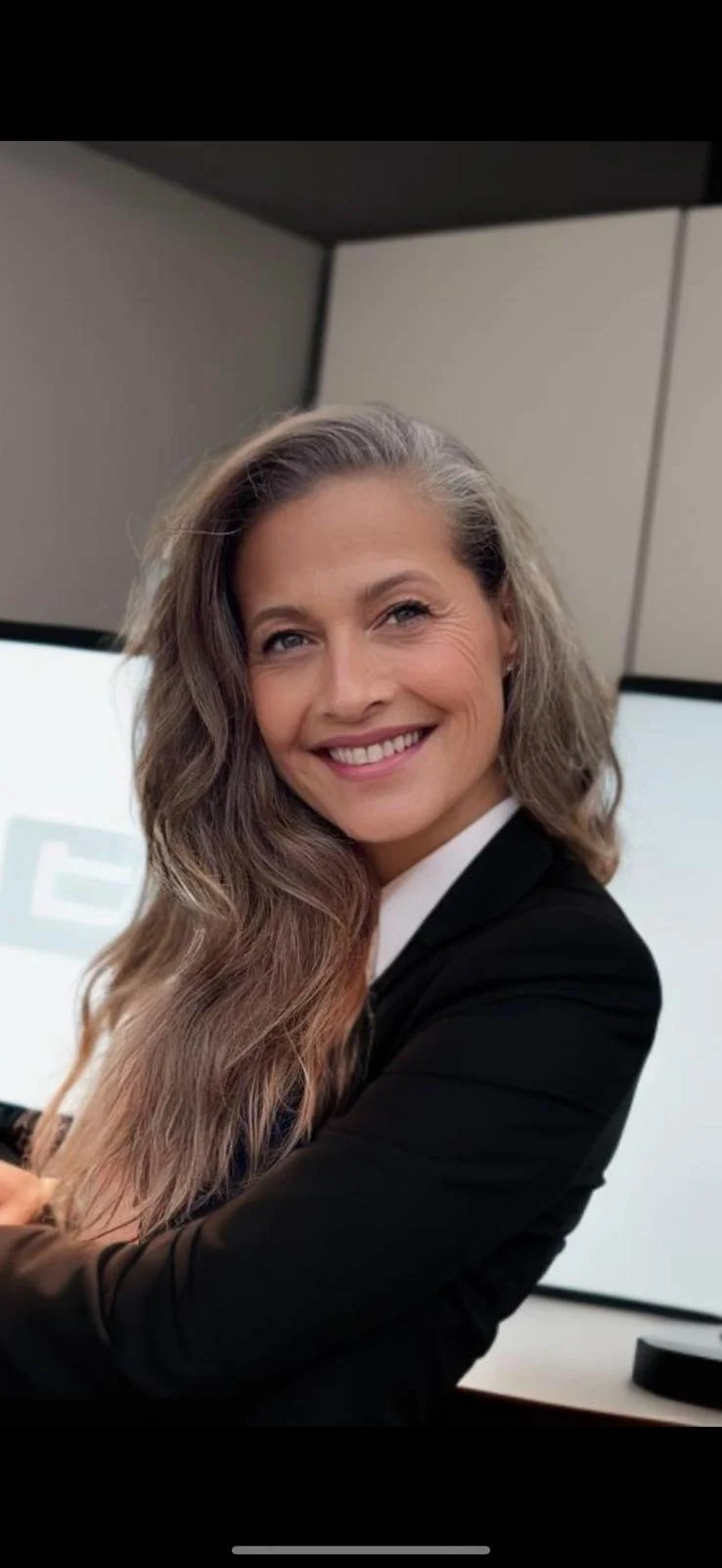 A woman with long, wavy, brown and gray hair smiling in a professional setting, wearing a black blazer over a white shirt, with a computer monitor in the background.