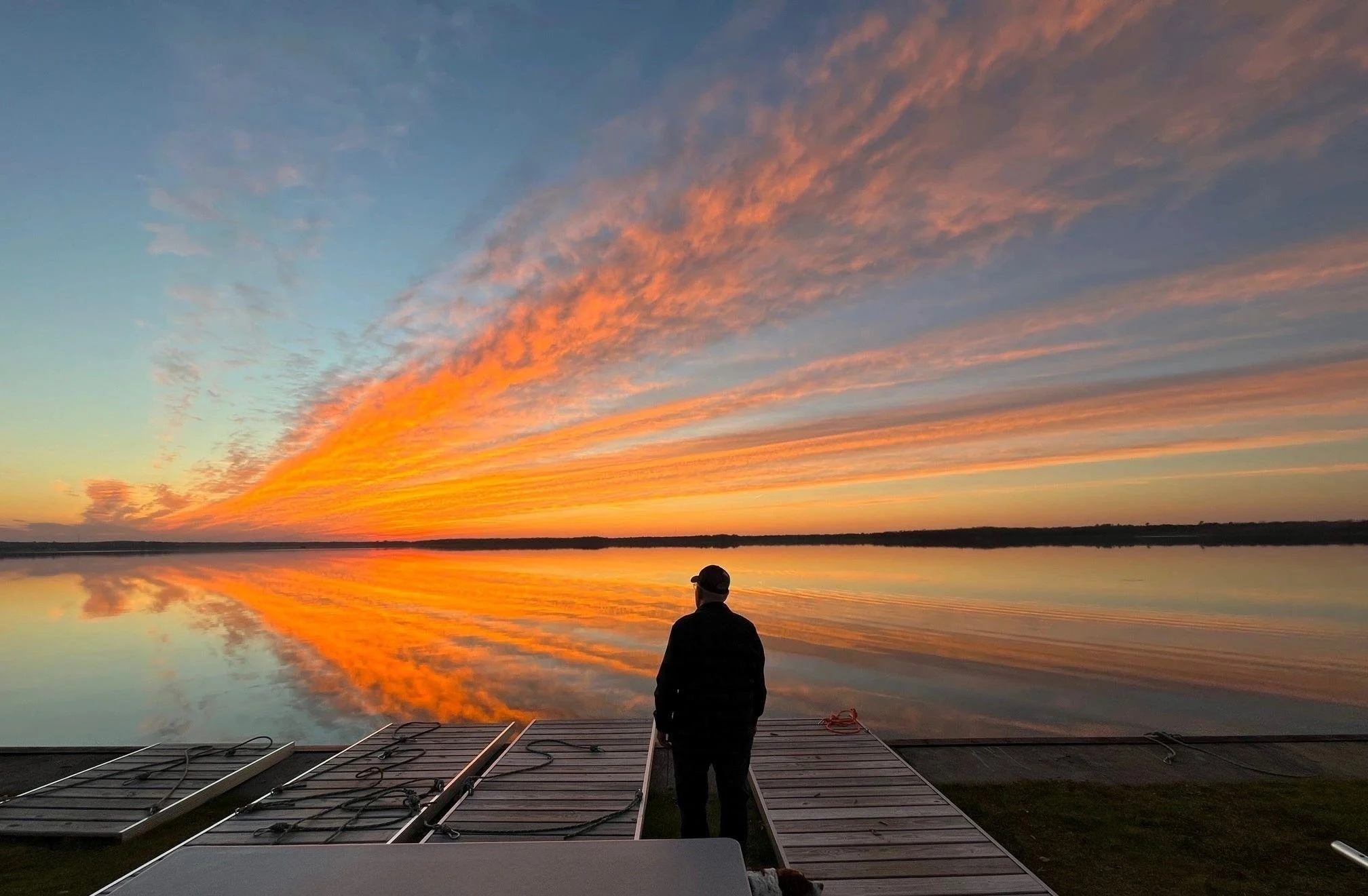 A person stands on a wooden dock at sunset, reflected in the calm water below.