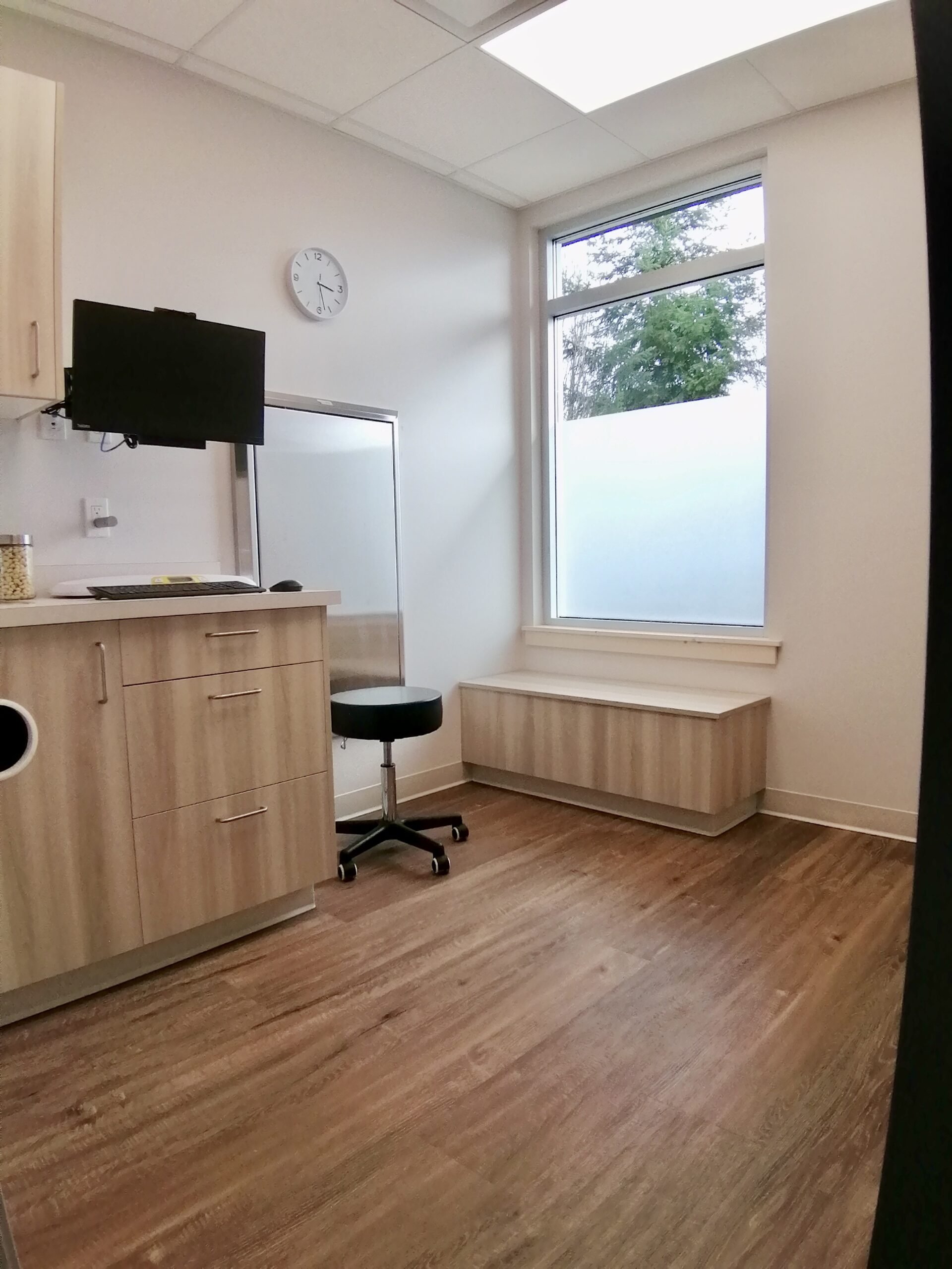 Empty room with wooden flooring, a large window with frosted glass, a wooden cabinet with a small television on top, a black office stool, and a white wall-mounted clock showing 1:11.