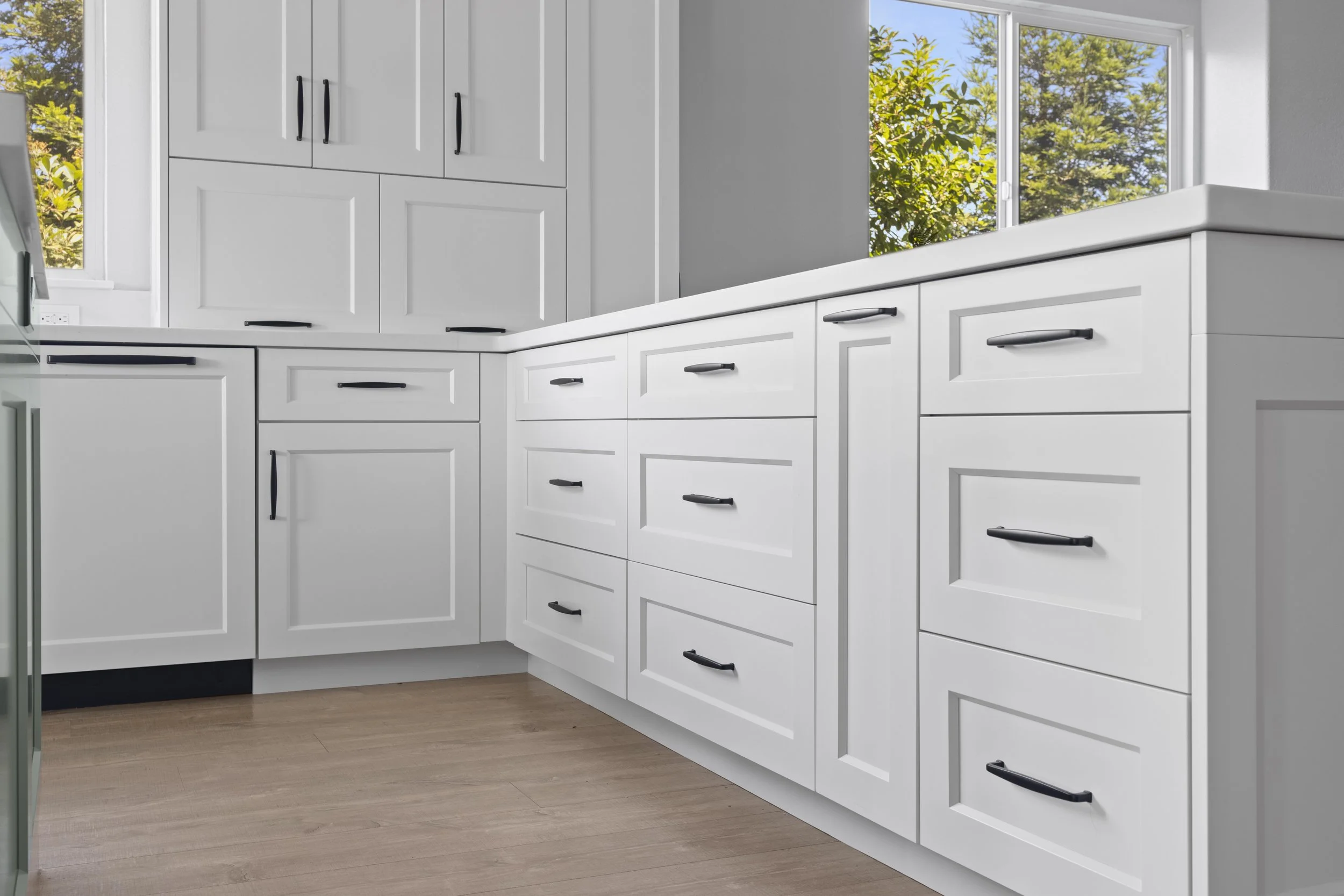Modern white kitchen cabinets with black handles, a window showing trees outside, and wooden flooring.