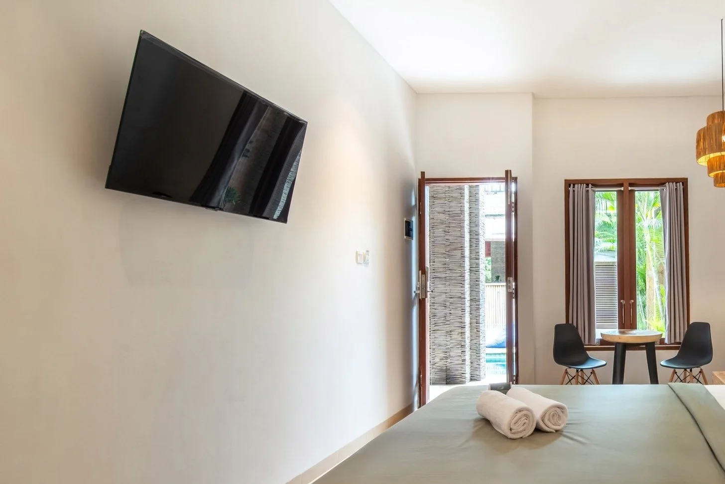 Modern hotel room with wall-mounted TV, bed with rolled towels, and small table with two chairs by window.