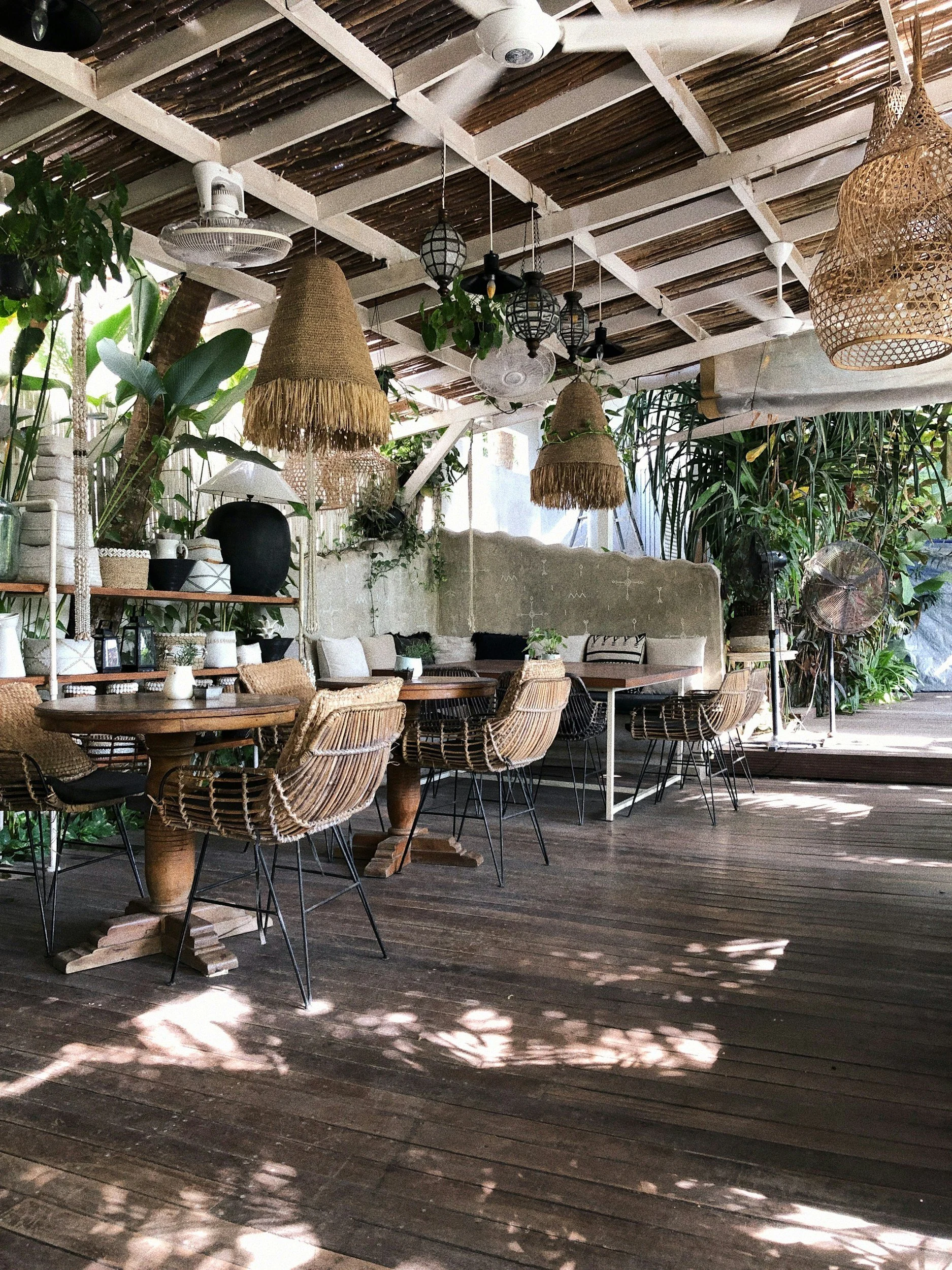 Outdoor patio with wicker furniture, wooden tables, thatched lampshades, ceiling fans, and greenery.