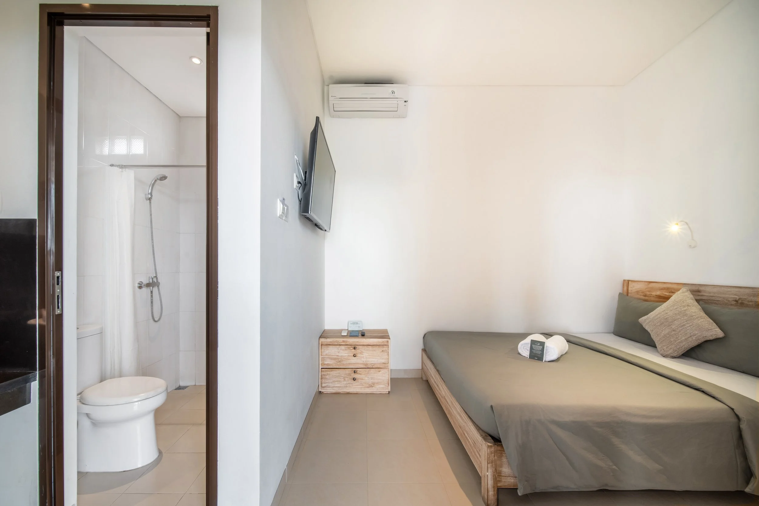 Minimalist hotel room with a wooden bed, gray bedding, wall-mounted TV, air conditioning, nightstand, and visible bathroom with toilet and shower.