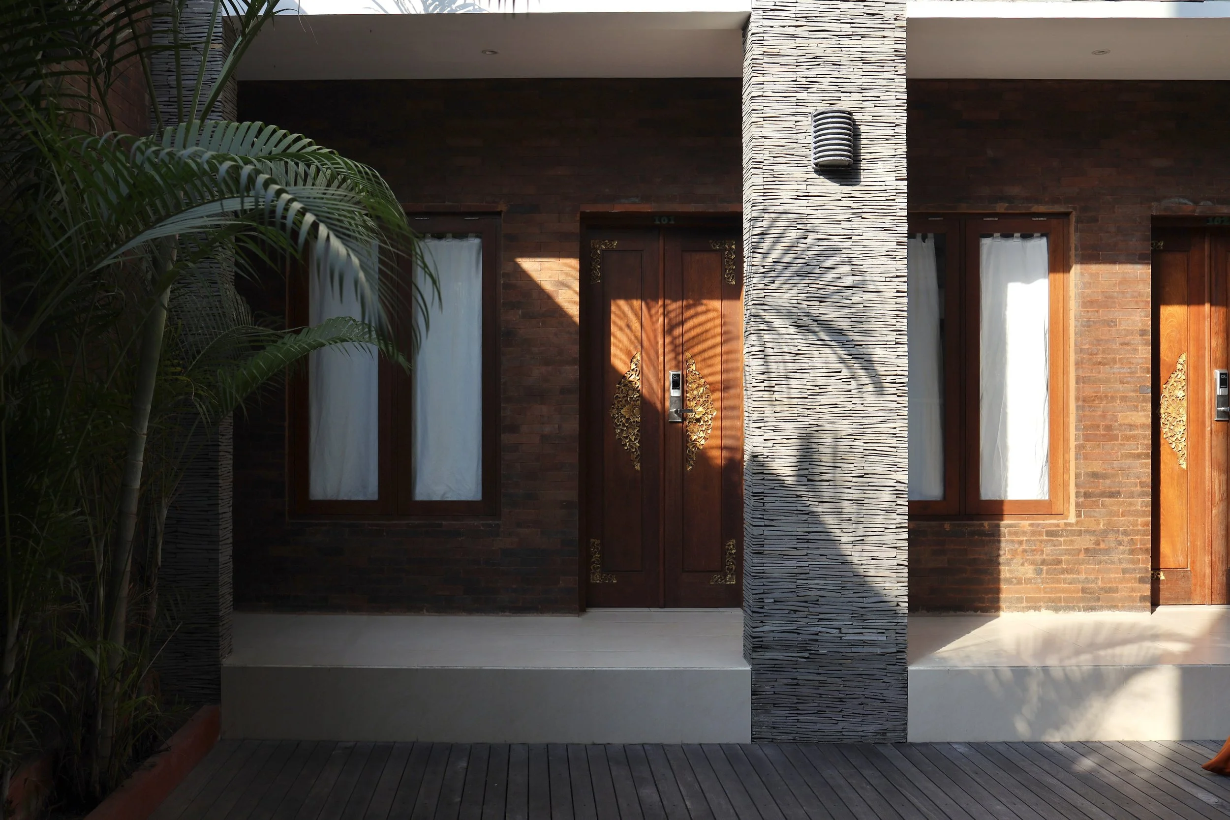 Entrance with wooden carved doors, brick walls, stone pillar, and palm leaves.