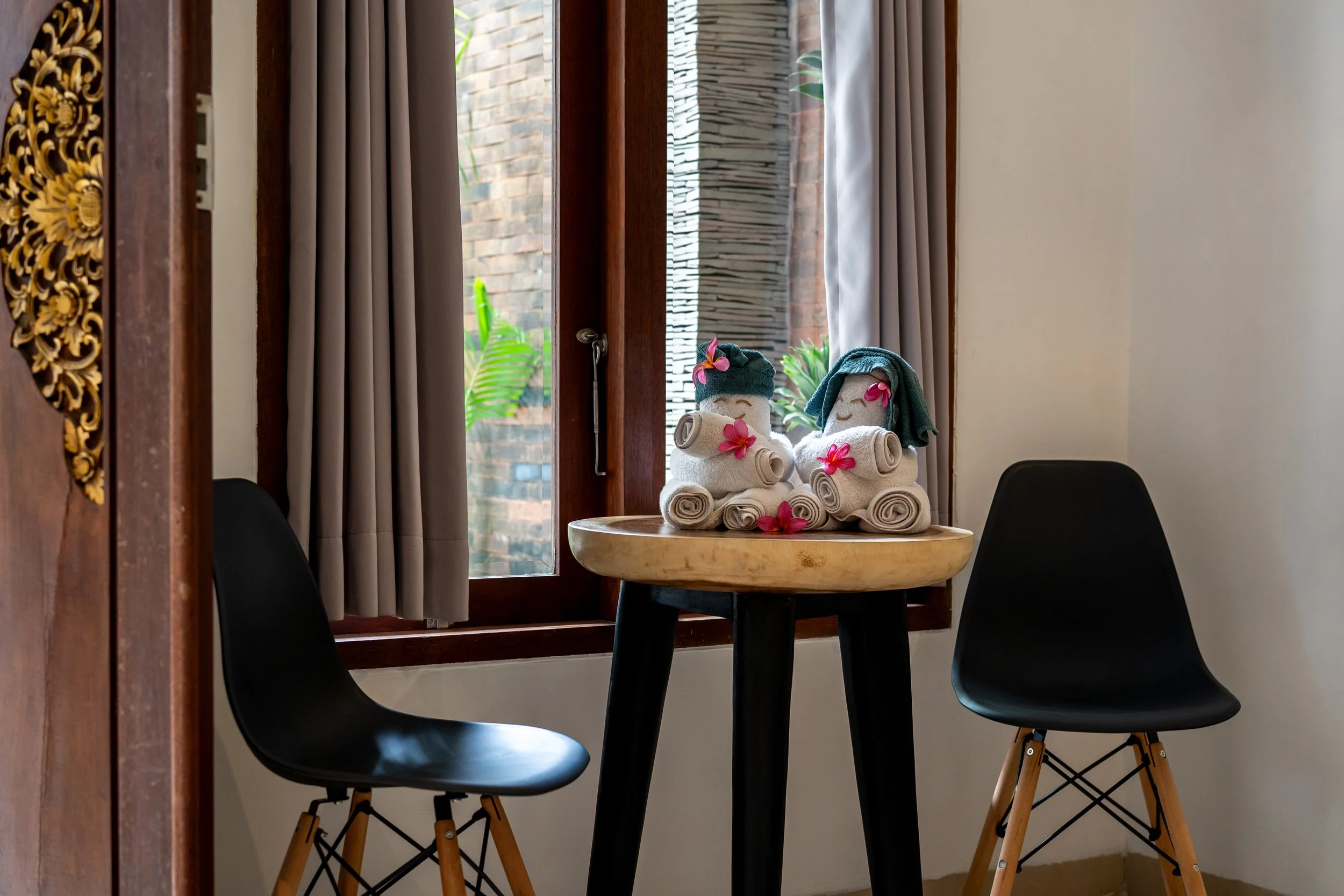 Cozy interior with two black chairs and a small wooden table holding two towel sculptures resembling teddy bears with flowers, near a window with curtains.
