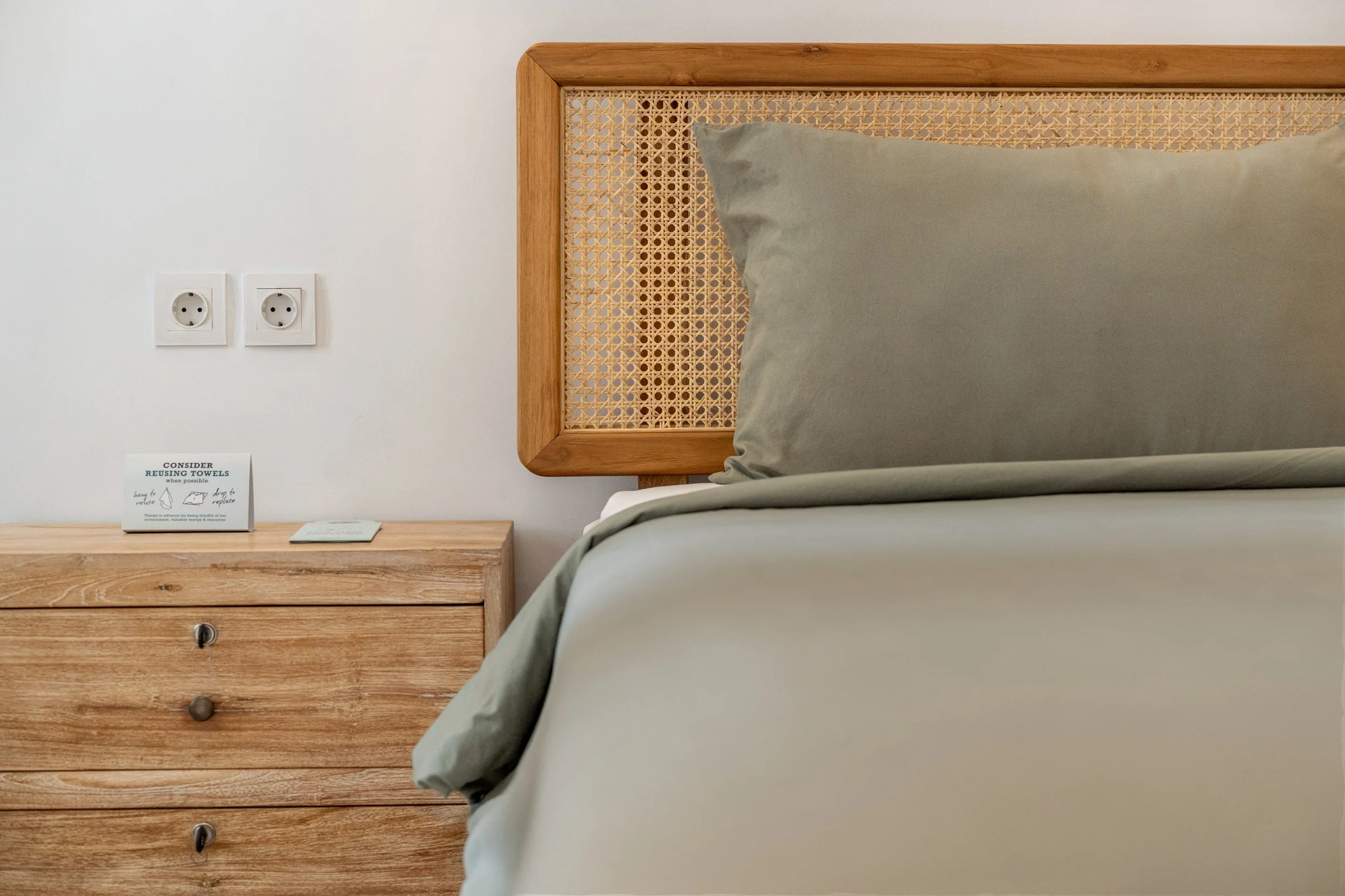 Hotel room with wooden headboard, gray pillow, and duvet, wooden bedside table, and electrical outlets on the wall.