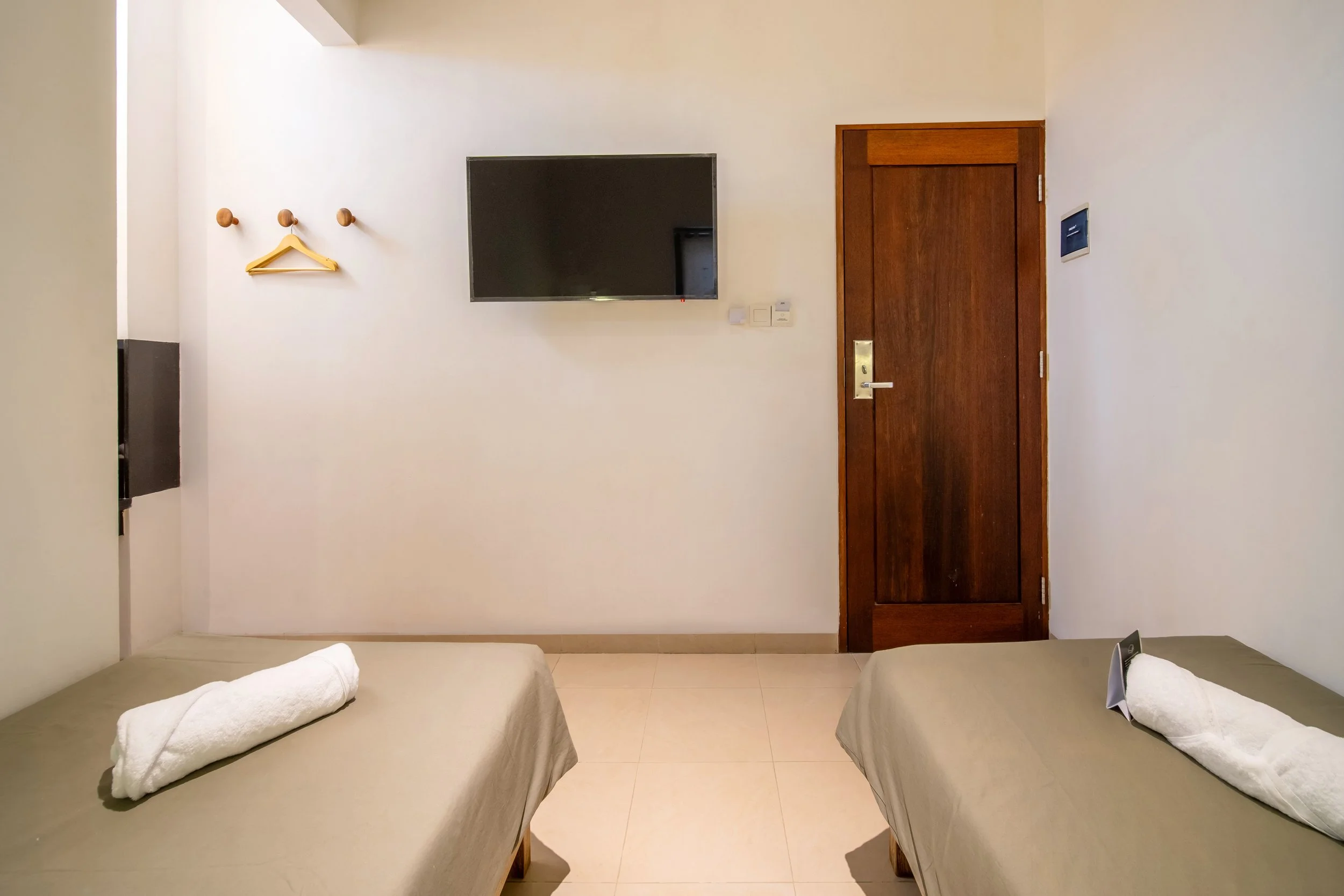 Minimalist hotel room with two beds, each with a towel, a wall-mounted TV, a wooden door, and coat hooks with a hanger.