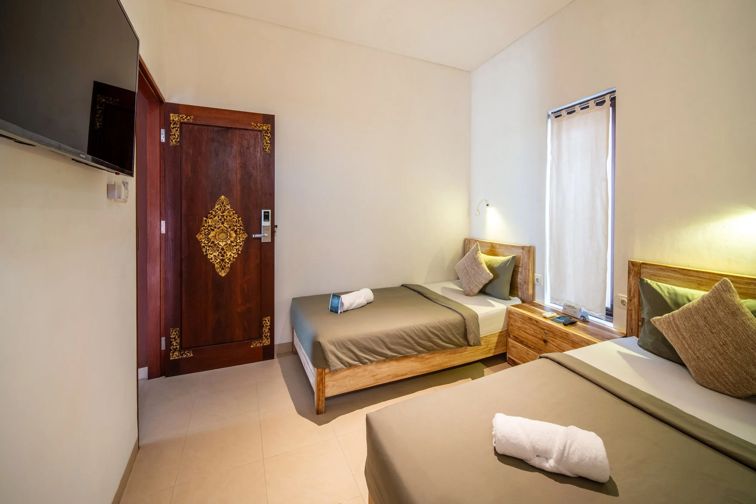 Hotel room with two single beds, wooden headboards, decorative pillow and towel on each, wall-mounted TV, ornate wooden door, closed window curtain, and bedside table.