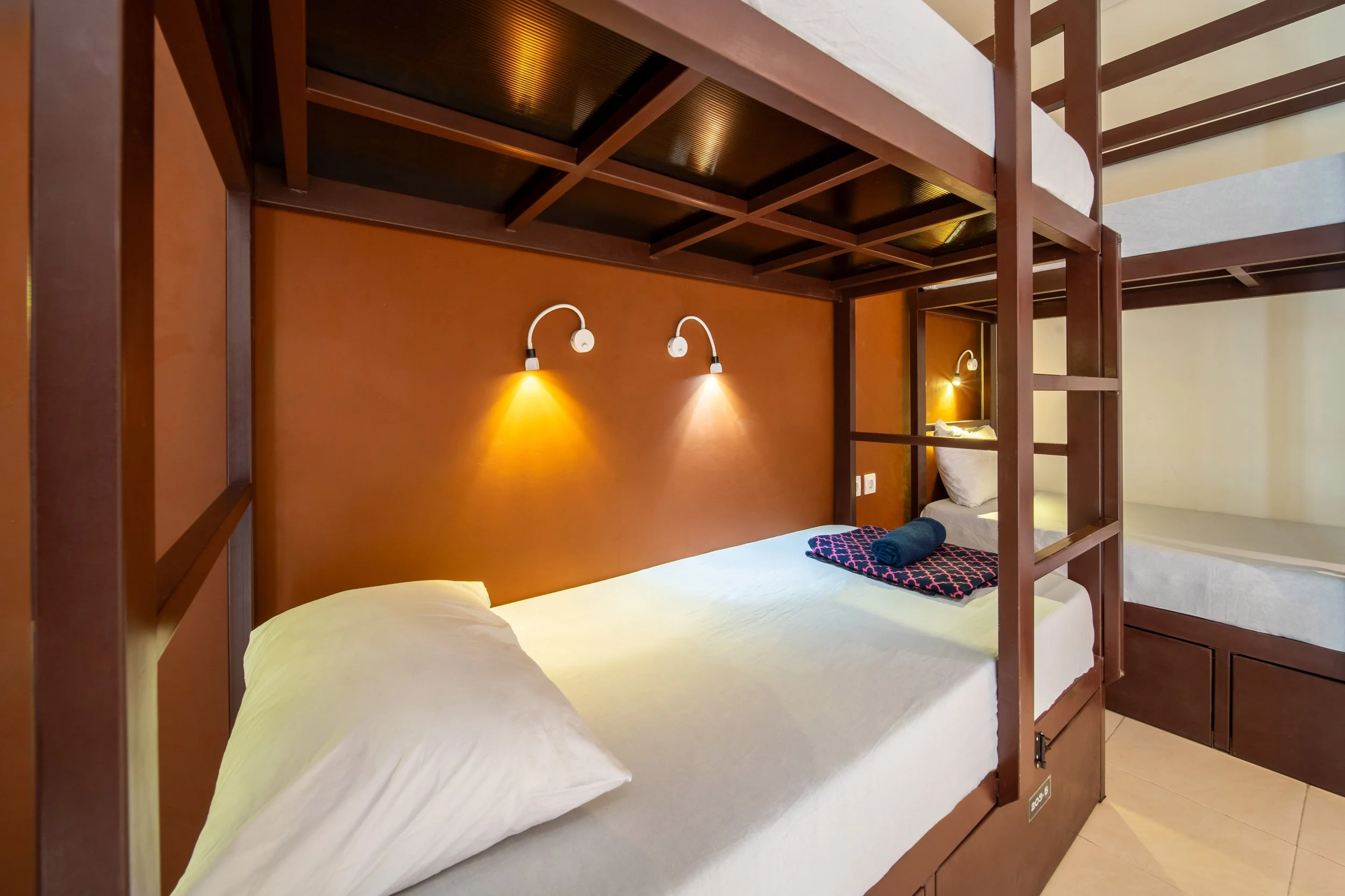 Two bunk beds with wooden frames and white bedding, featuring individual reading lights on the wall and folded towels placed on one of the beds.