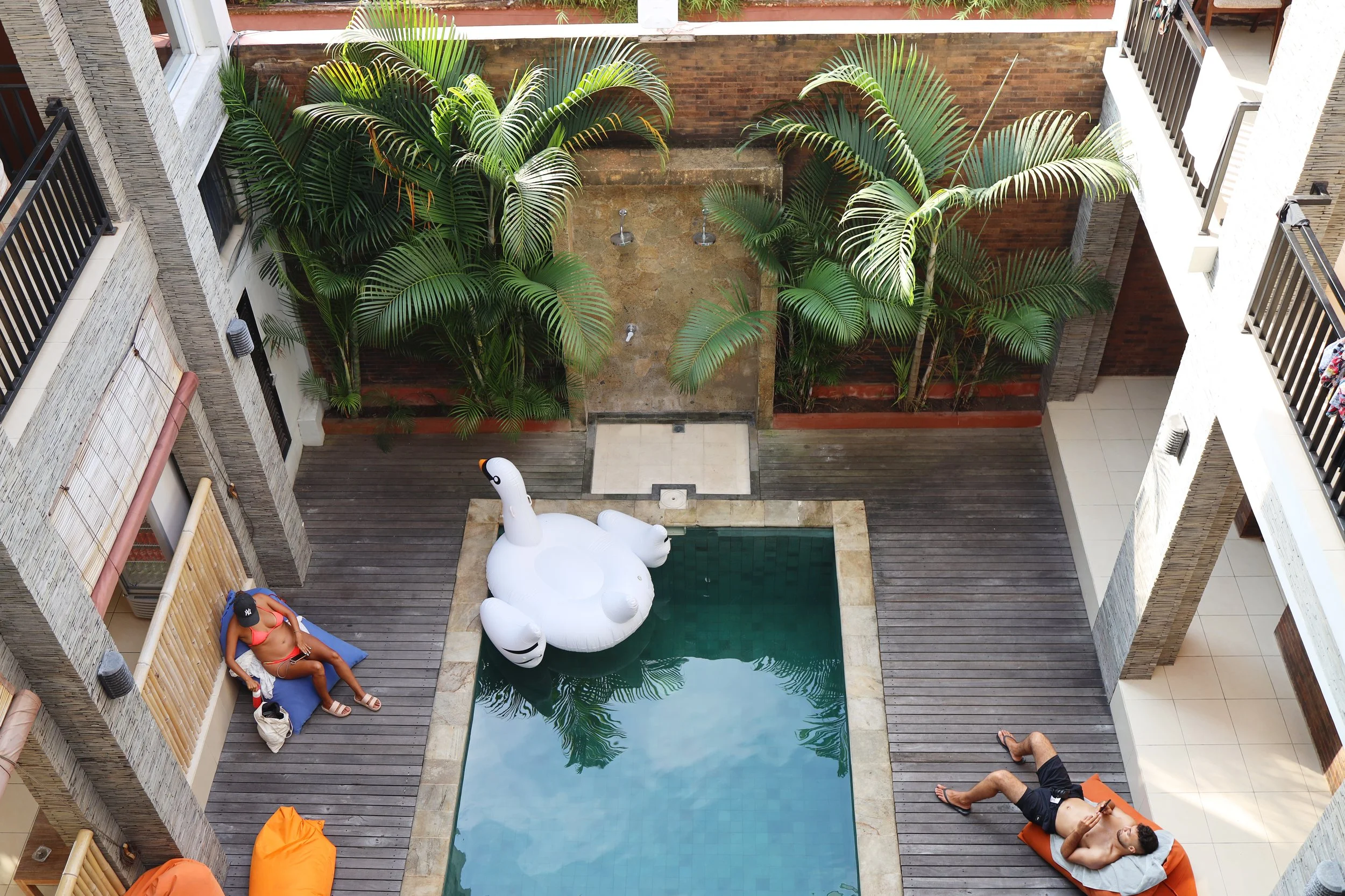 Aerial view of a small swimming pool with a white inflatable swan float. Two people are sunbathing on loungers, surrounded by tropical plants and wooden decking.