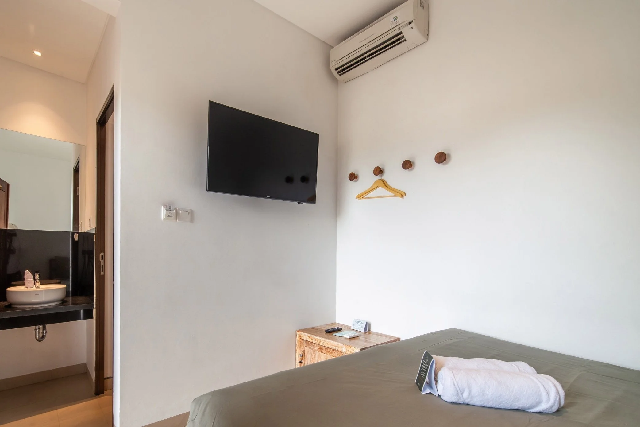 Hotel room interior with air conditioner, wall-mounted TV, wooden hangers, rolled towel on bed, and a view of a bathroom sink.
