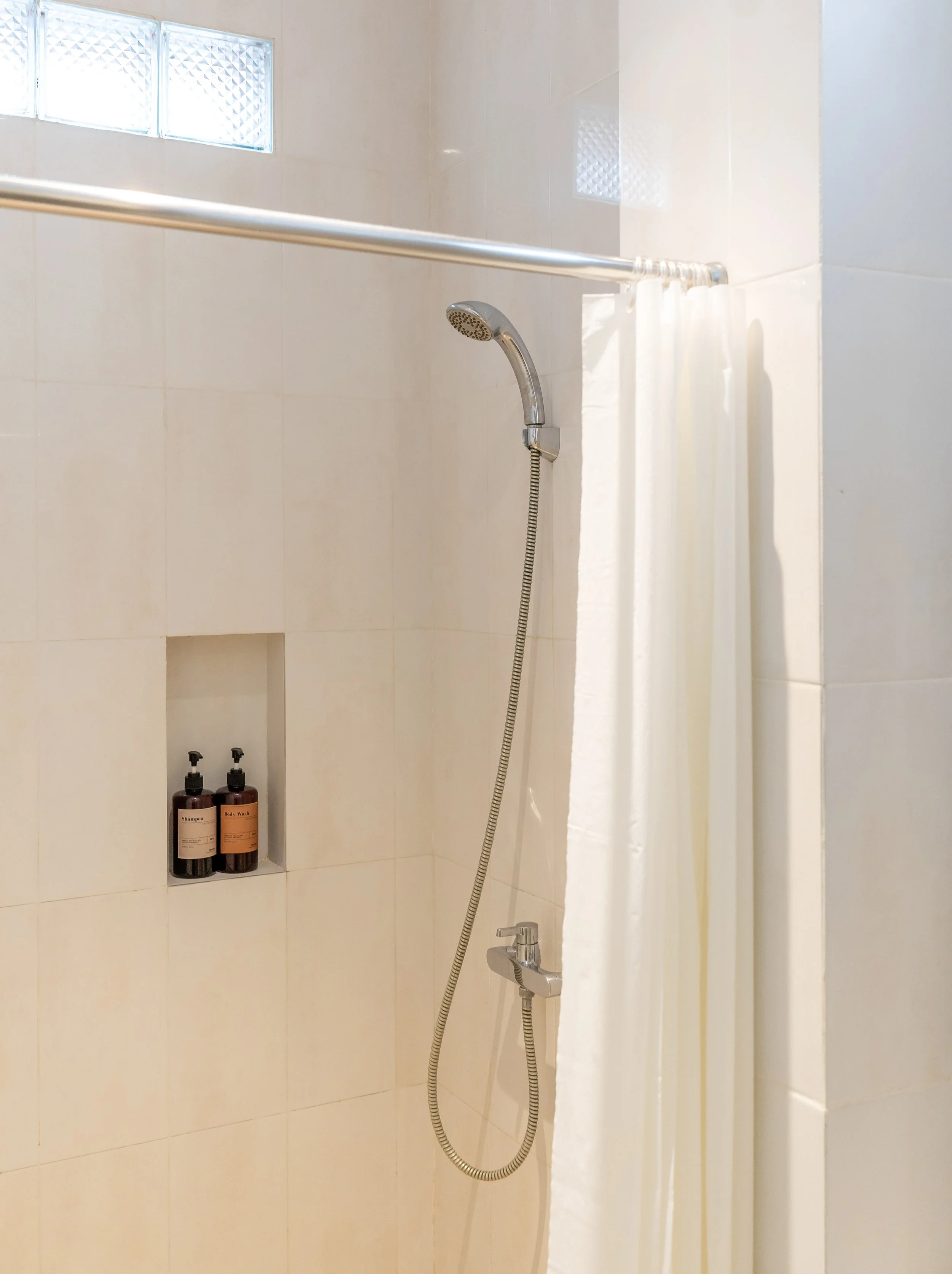 Modern bathroom shower with a showerhead, a white shower curtain, and two bottles of bath products on a built-in shelf.