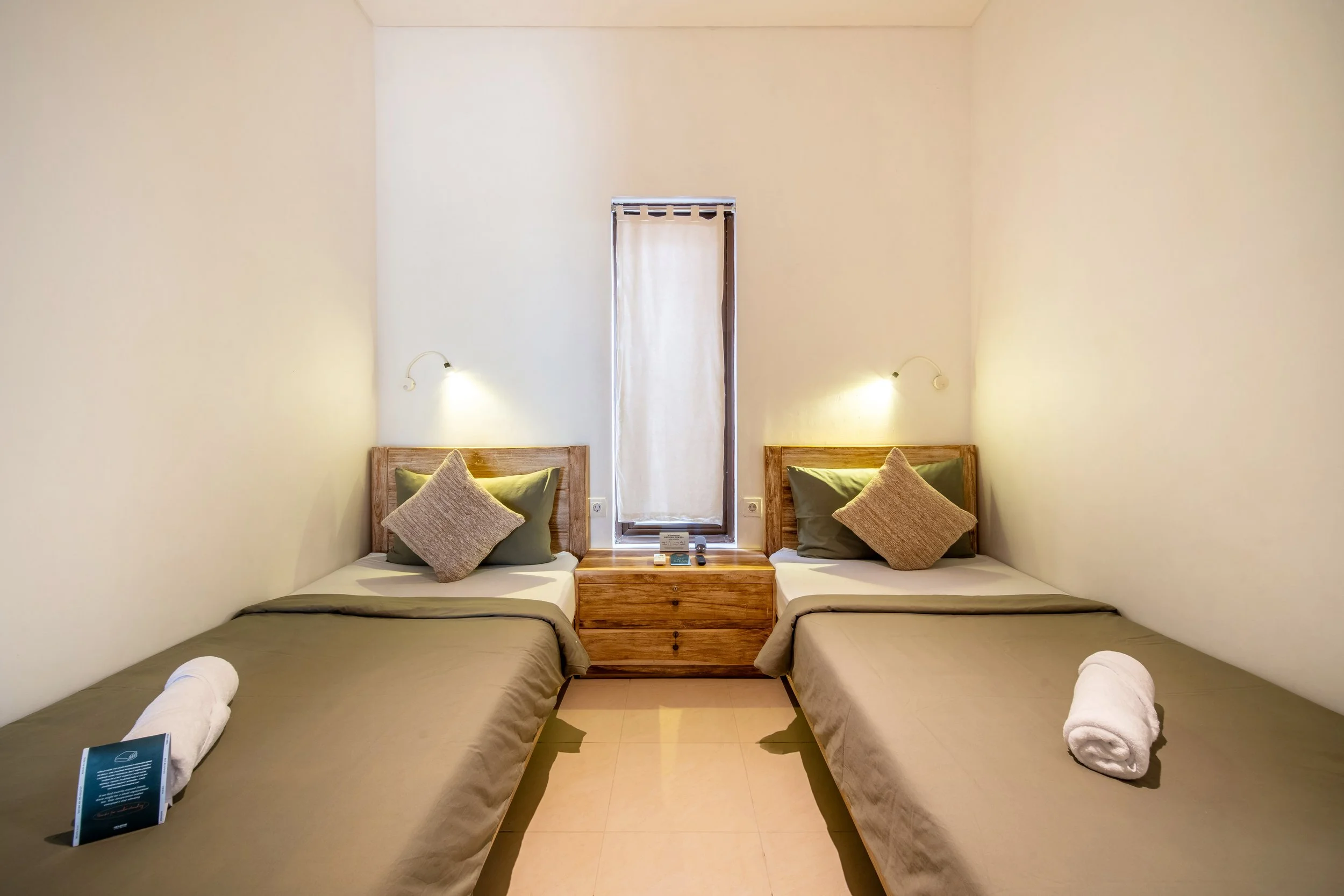 Minimalist bedroom with two twin beds, wooden headboards, beige and green pillows, bedside table, wall-mounted lamps, and a narrow window with a curtain.