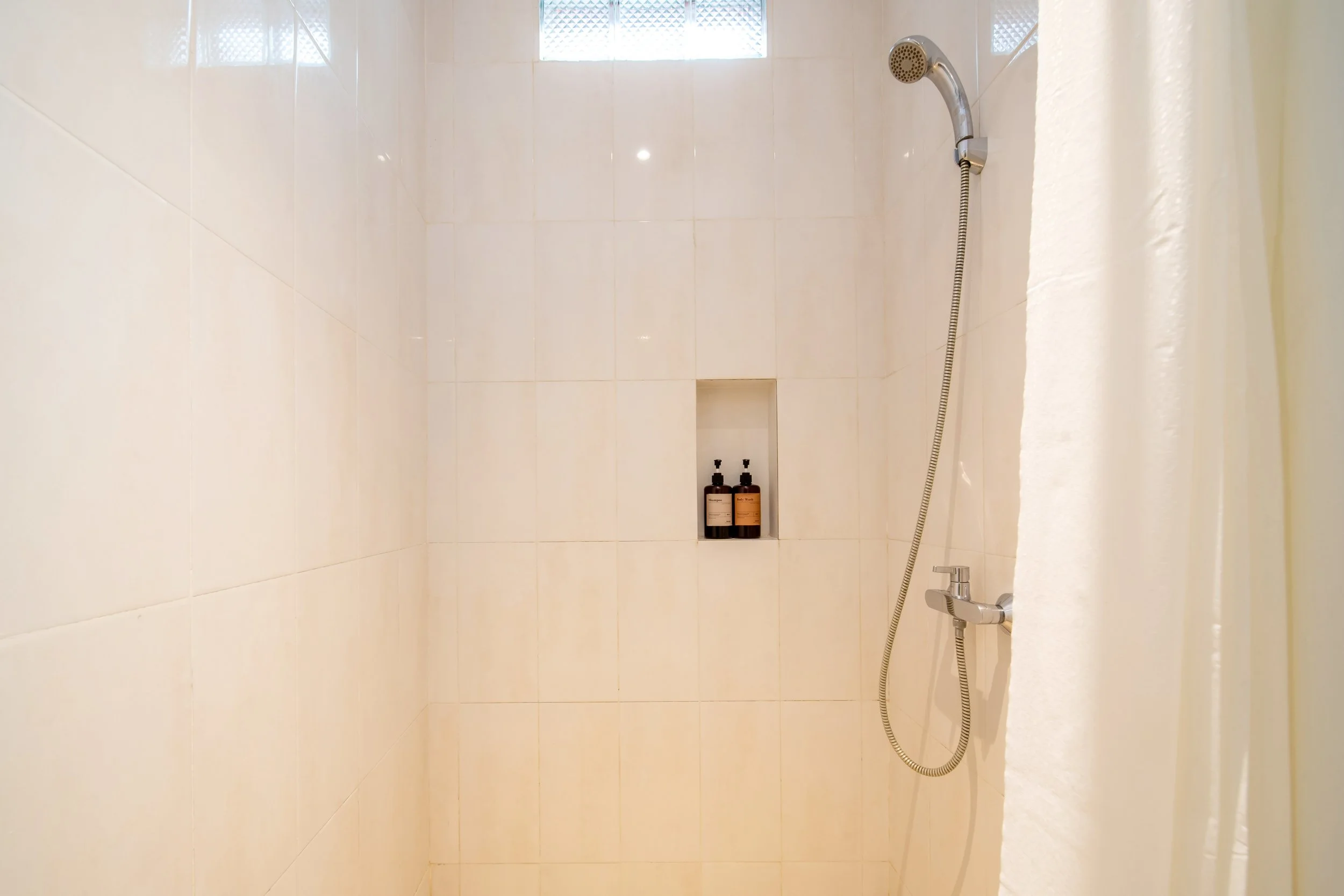 Minimalist shower with beige tiles, handheld showerhead, and two bottles in a recessed shelf.