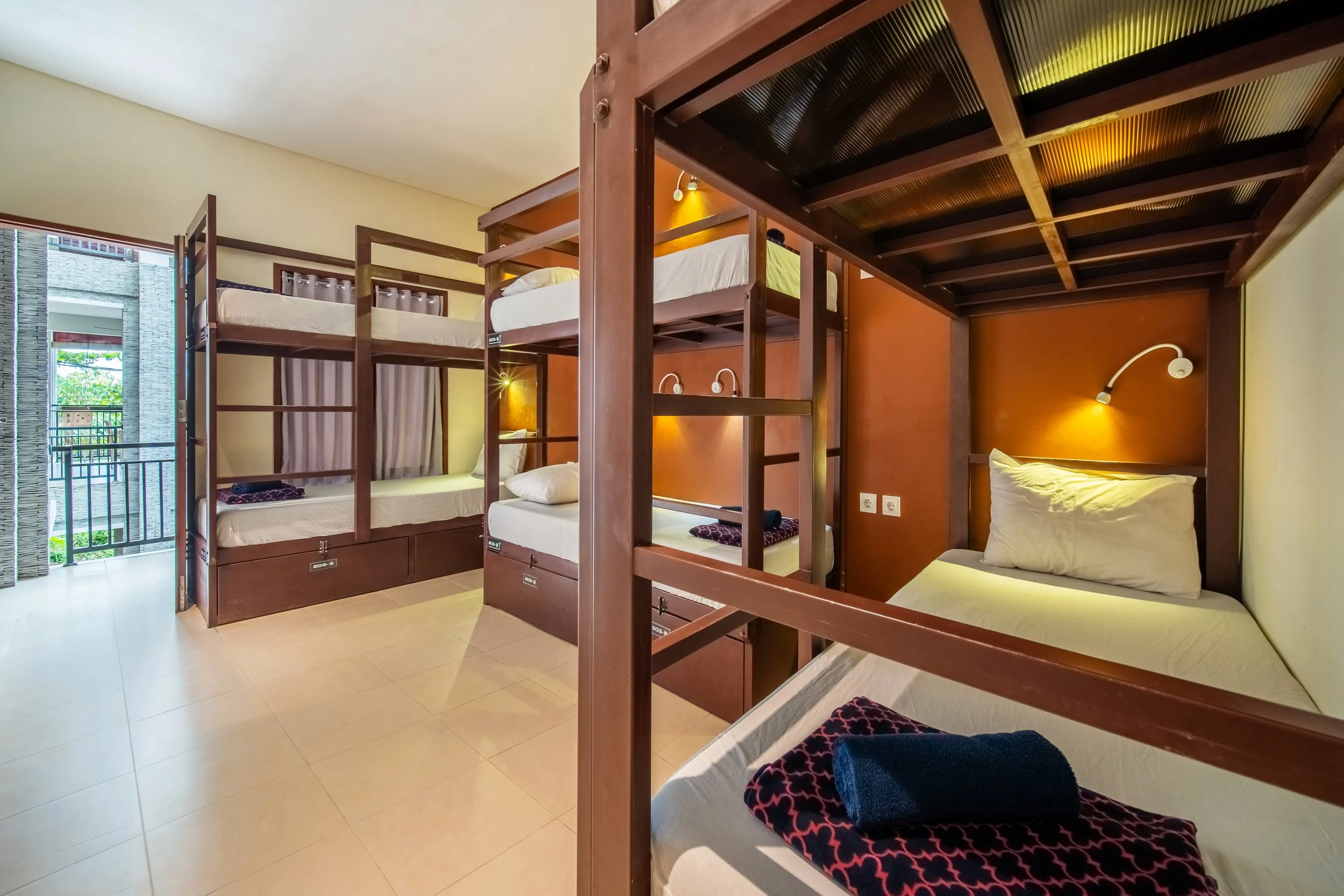 Room with four bunk beds, individual reading lights, and neatly folded towels on each bed. A balcony with greenery is visible through an open door.