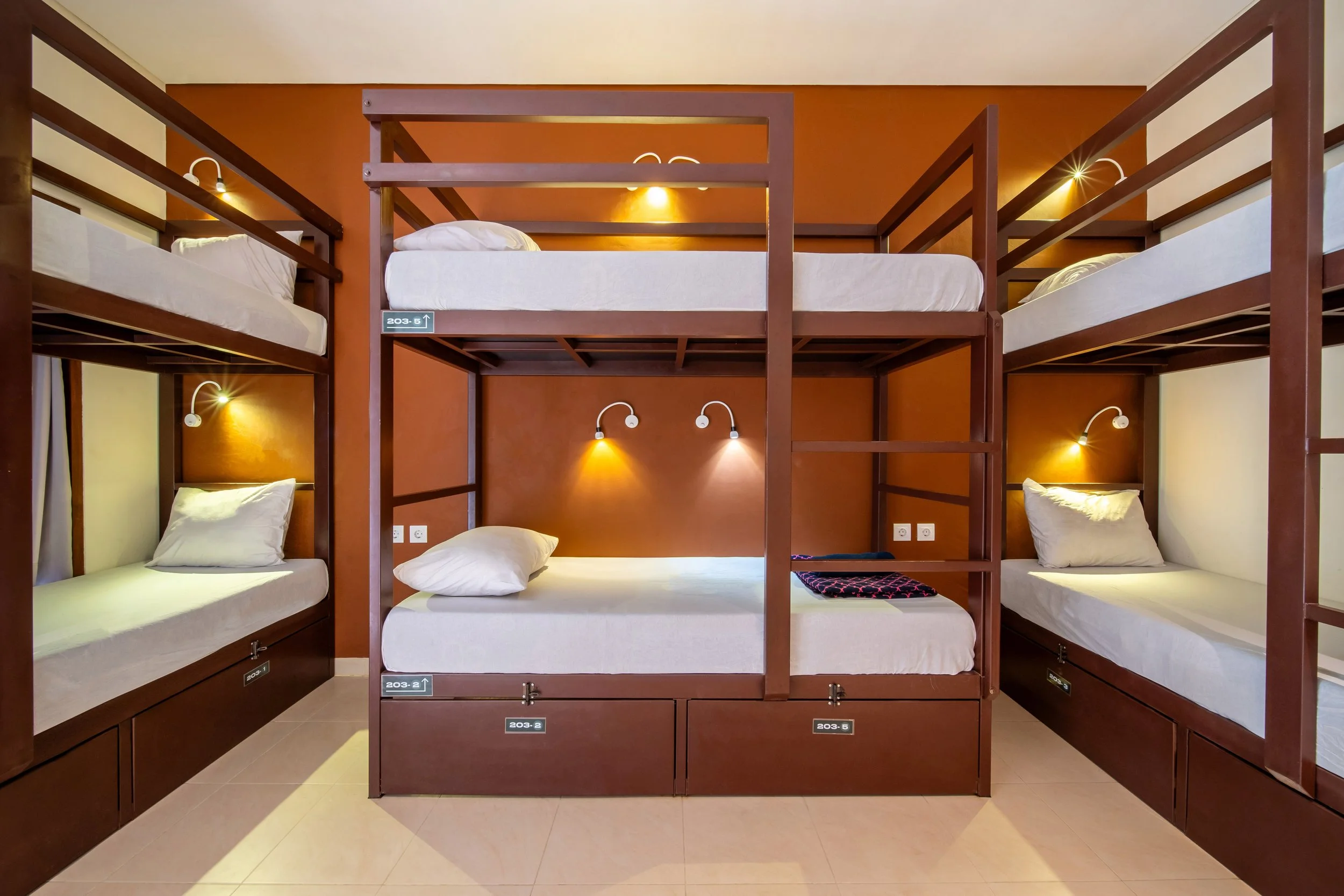 A dormitory room with wooden bunk beds, white bedding, and wall-mounted reading lights.
