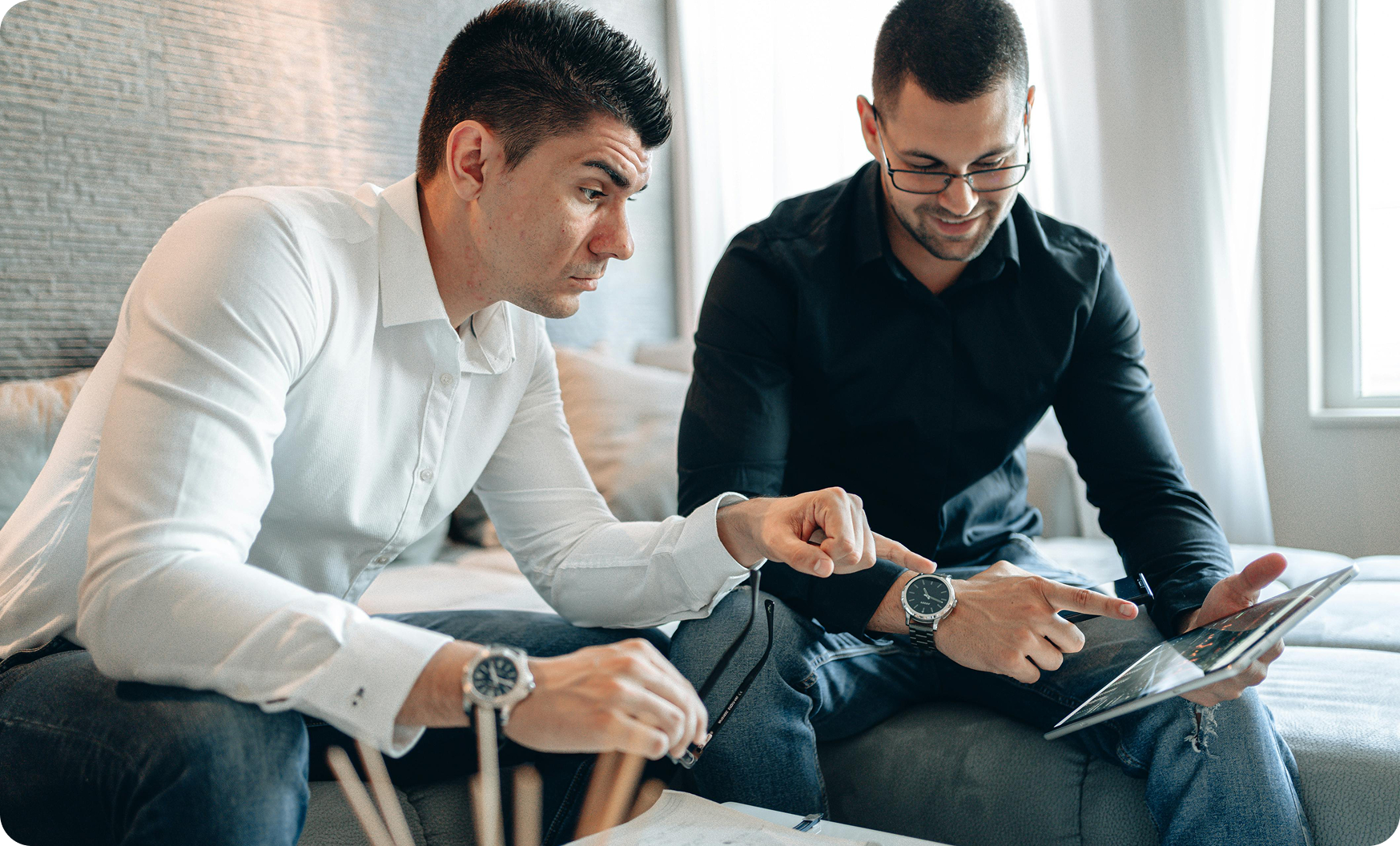 Two men sitting indoors, one in a white shirt holding glasses, pointing at a tablet held by the other man in a black shirt.