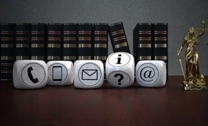 Row of six decorative cubes with icons for phone, computer, email, information, question mark, and at symbol, placed in front of a row of black books, with a gold statue of Lady Justice on the right.