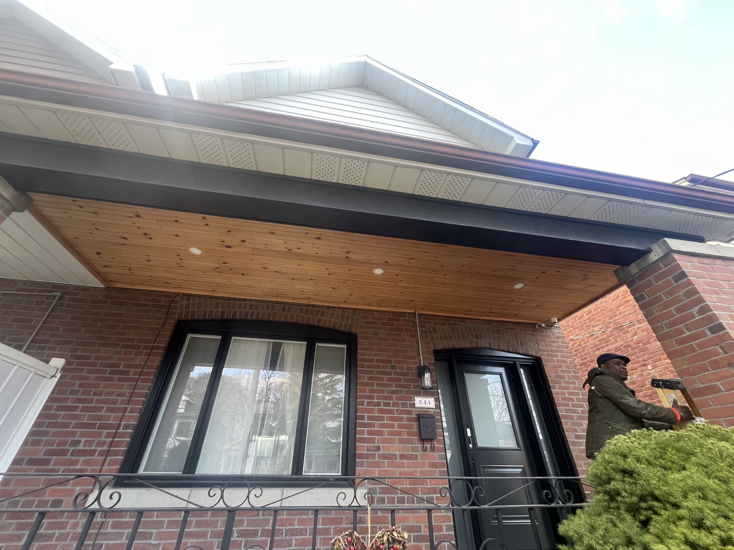 Construction worker installing a wooden ceiling on the porch of a brick house.