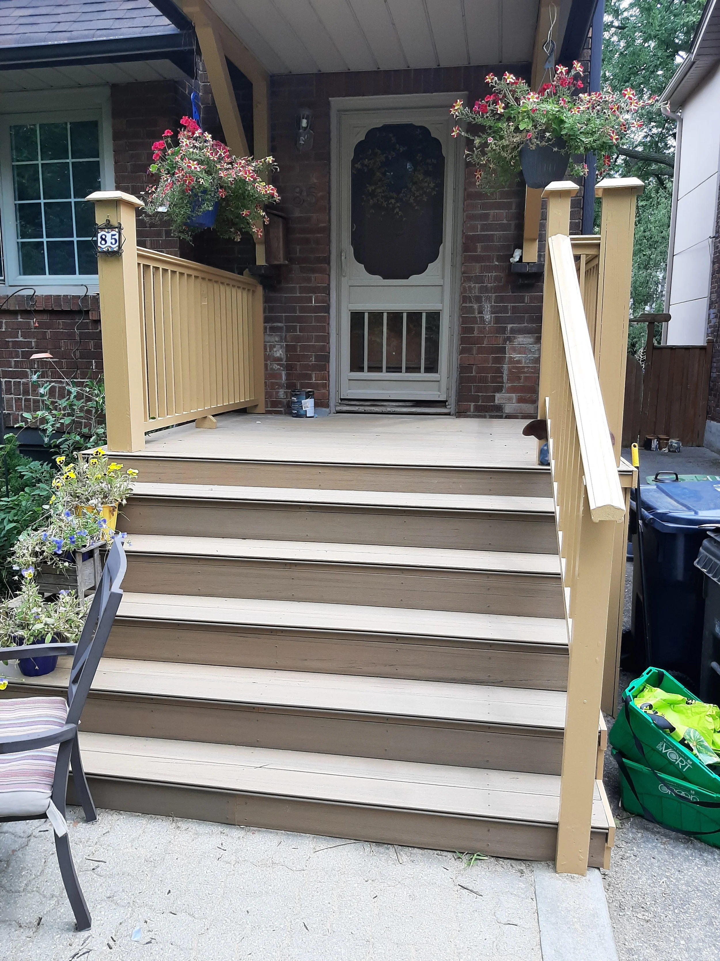 Newly built wooden front porch and stairs with beige railings and flower pots, in front of a brick house with a decorated door and blooming plants.