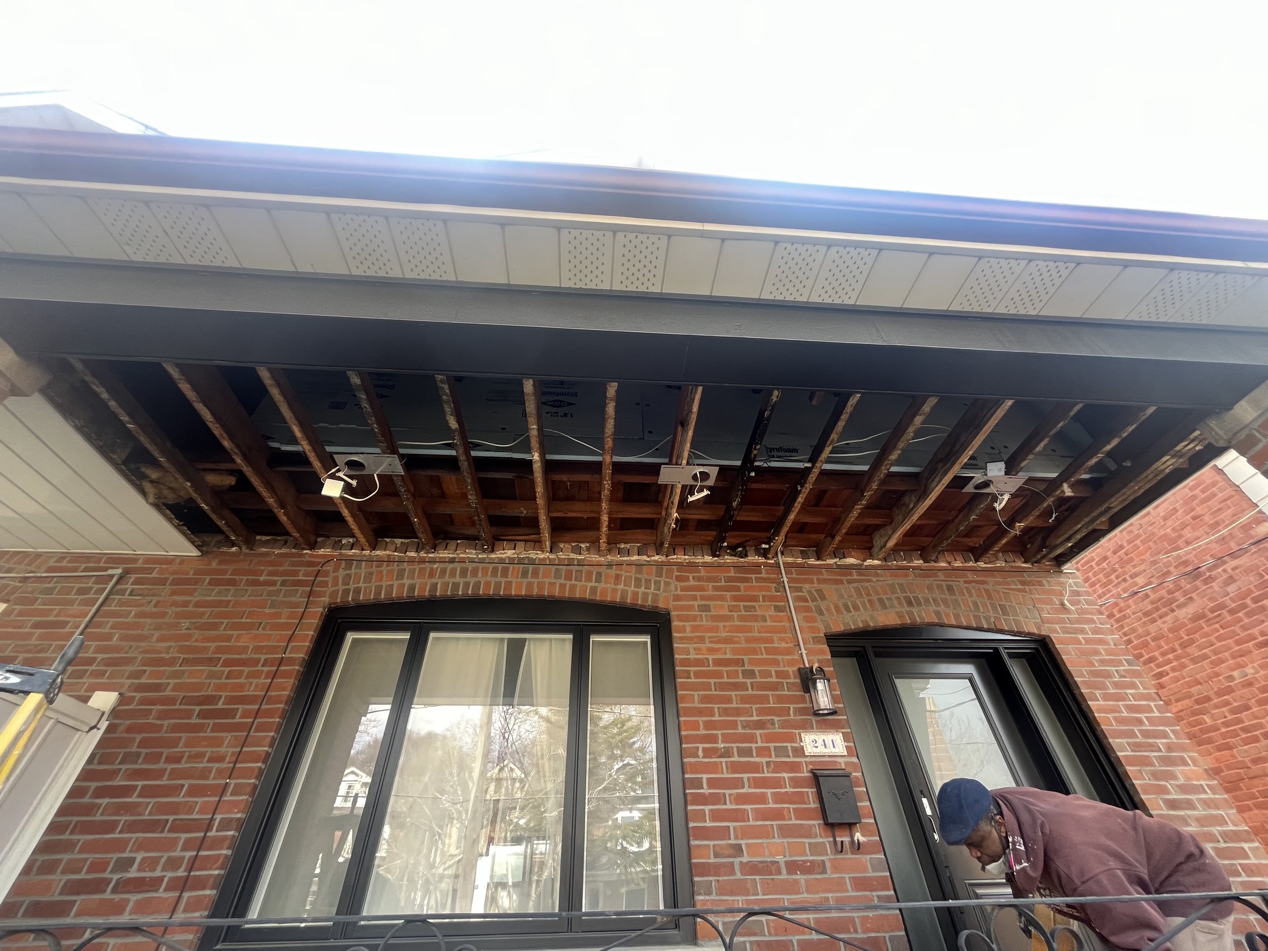 A man working on the exterior of a brick house, installing or repairing a ceiling or roof under the overhang, with exposed wooden beams and electrical wiring visible.