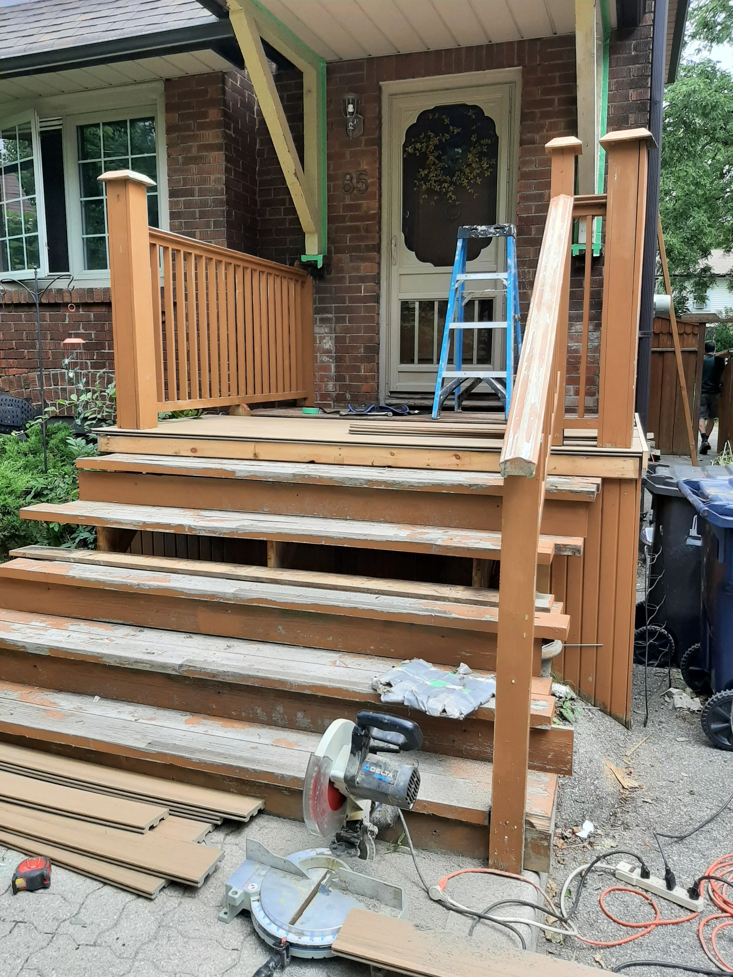 Front porch under renovation with new wooden railing and steps, construction tools including a miter saw and ladder, and materials scattered around.