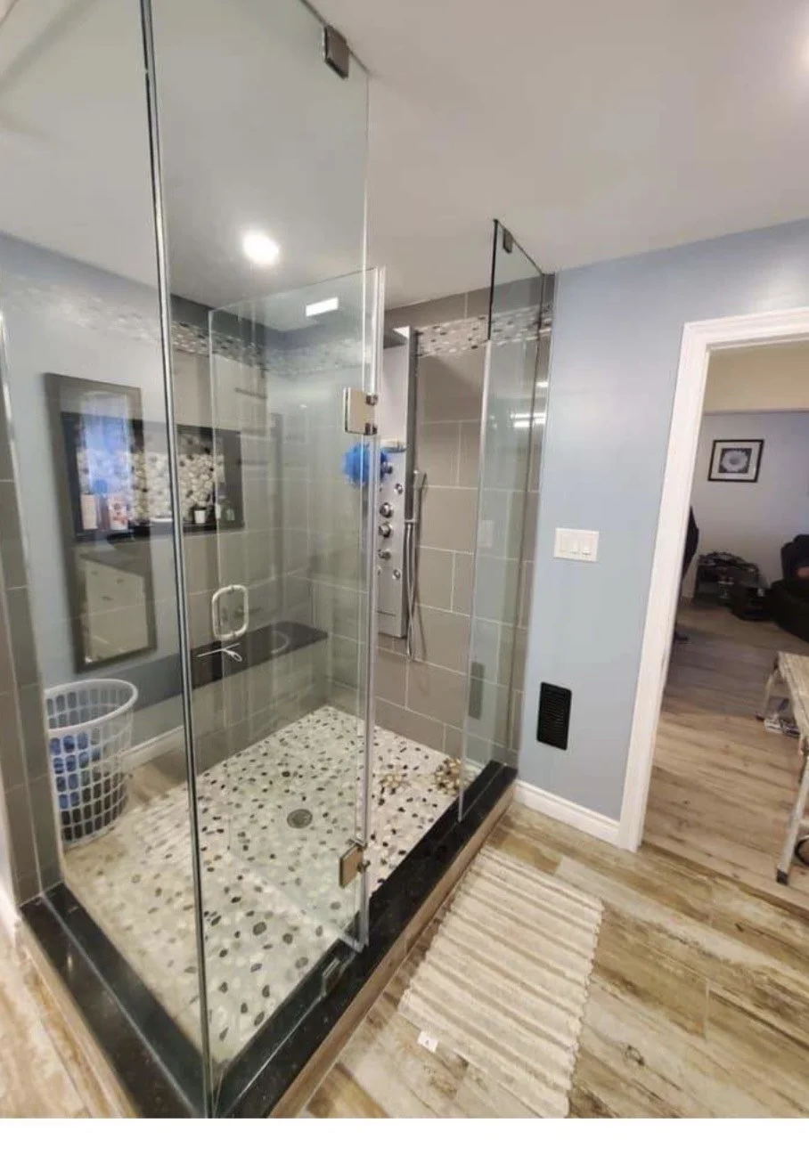 A modern glass shower with multicolored pebble tile floor, located in a room with hardwood floors and a rug outside.