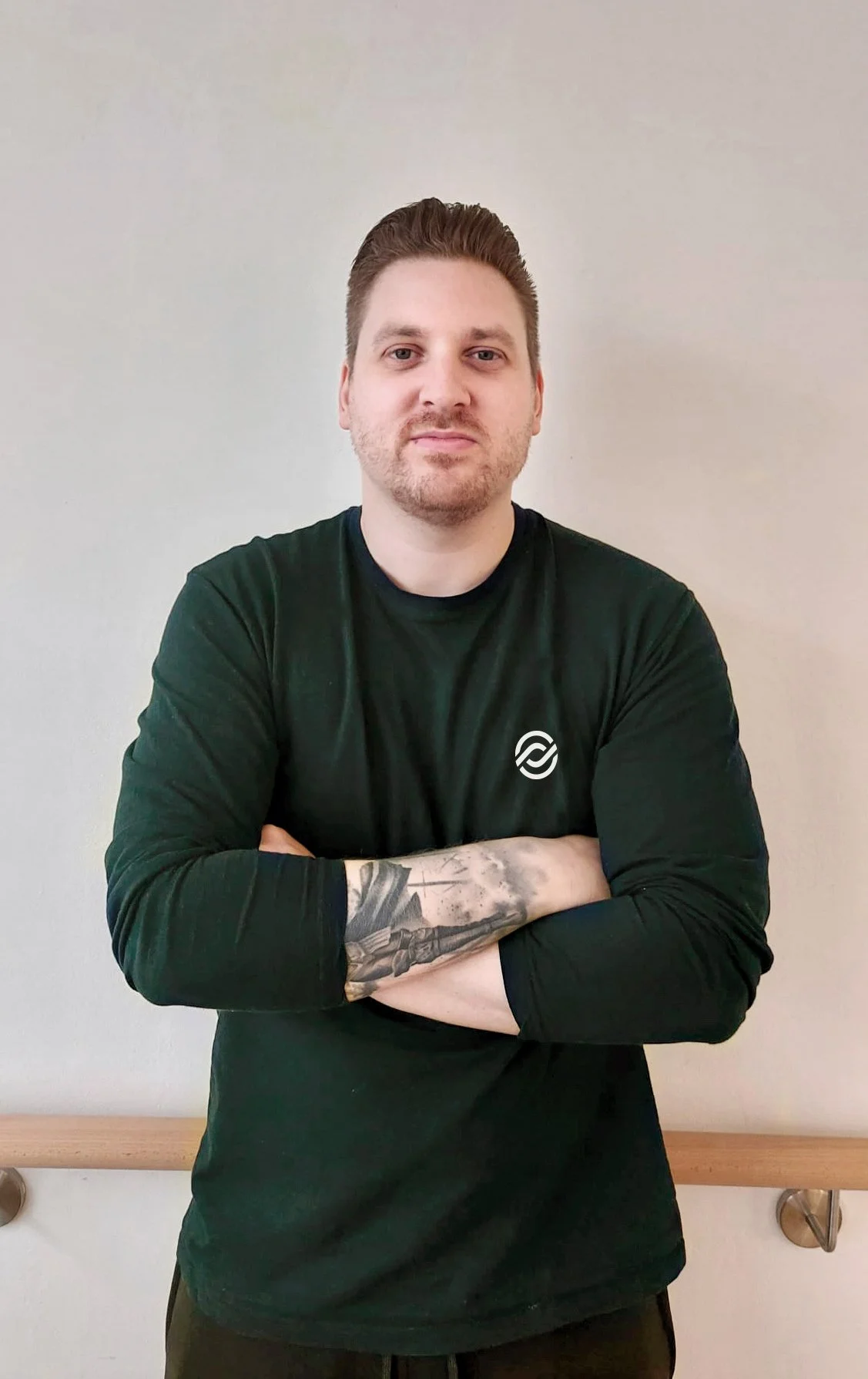 Ein Mann mit kurzen braunen Haaren, Bart und Tattoos trägt ein schwarzes T-Shirt mit einem weißen Logo, steht mit verschränkten Armen vor einer weißen Wand.