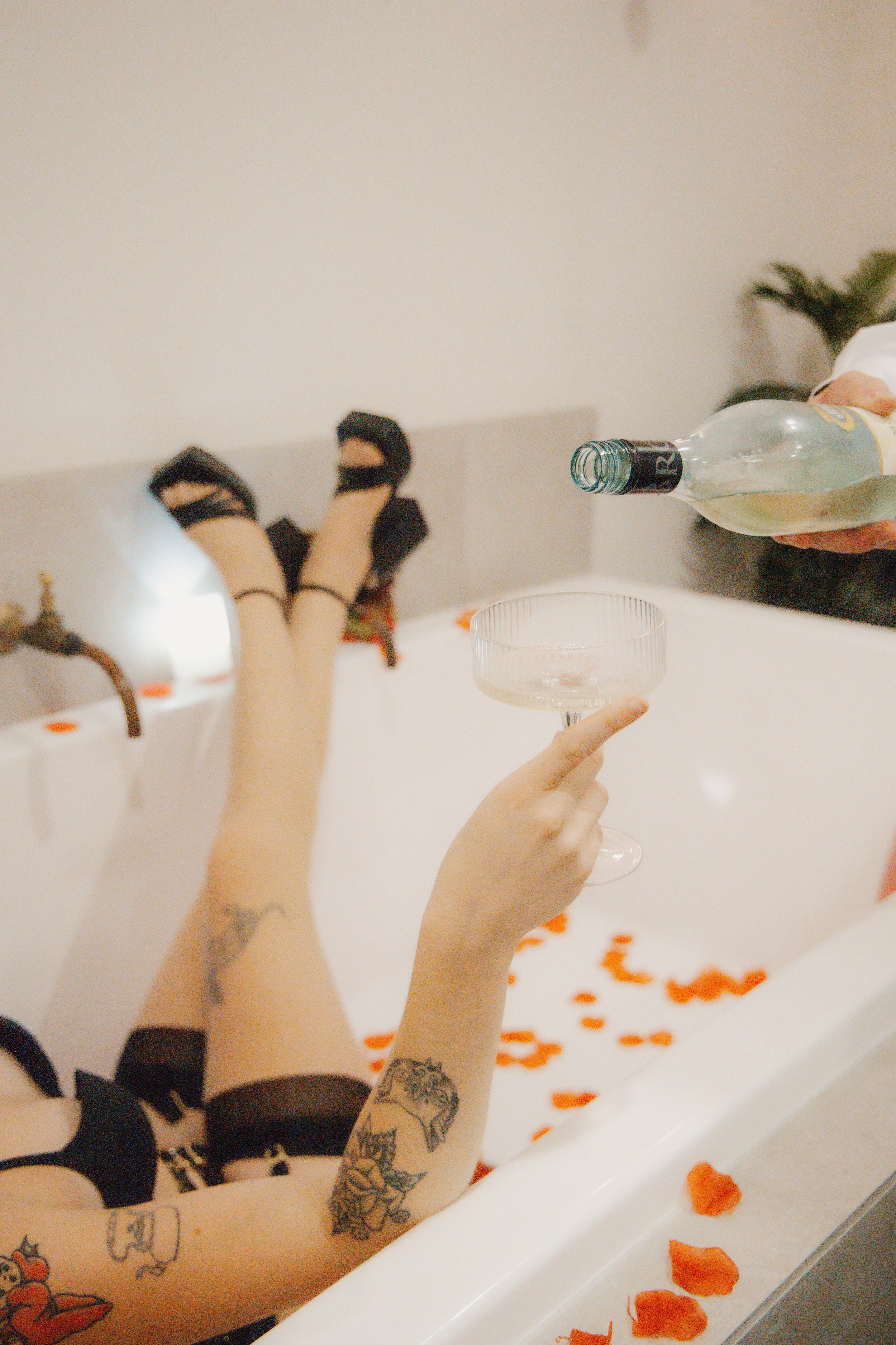 Person wearing black gloves and black shoes relaxing in a bathtub filled with rose petals, holding a glass and a bottle of white wine.