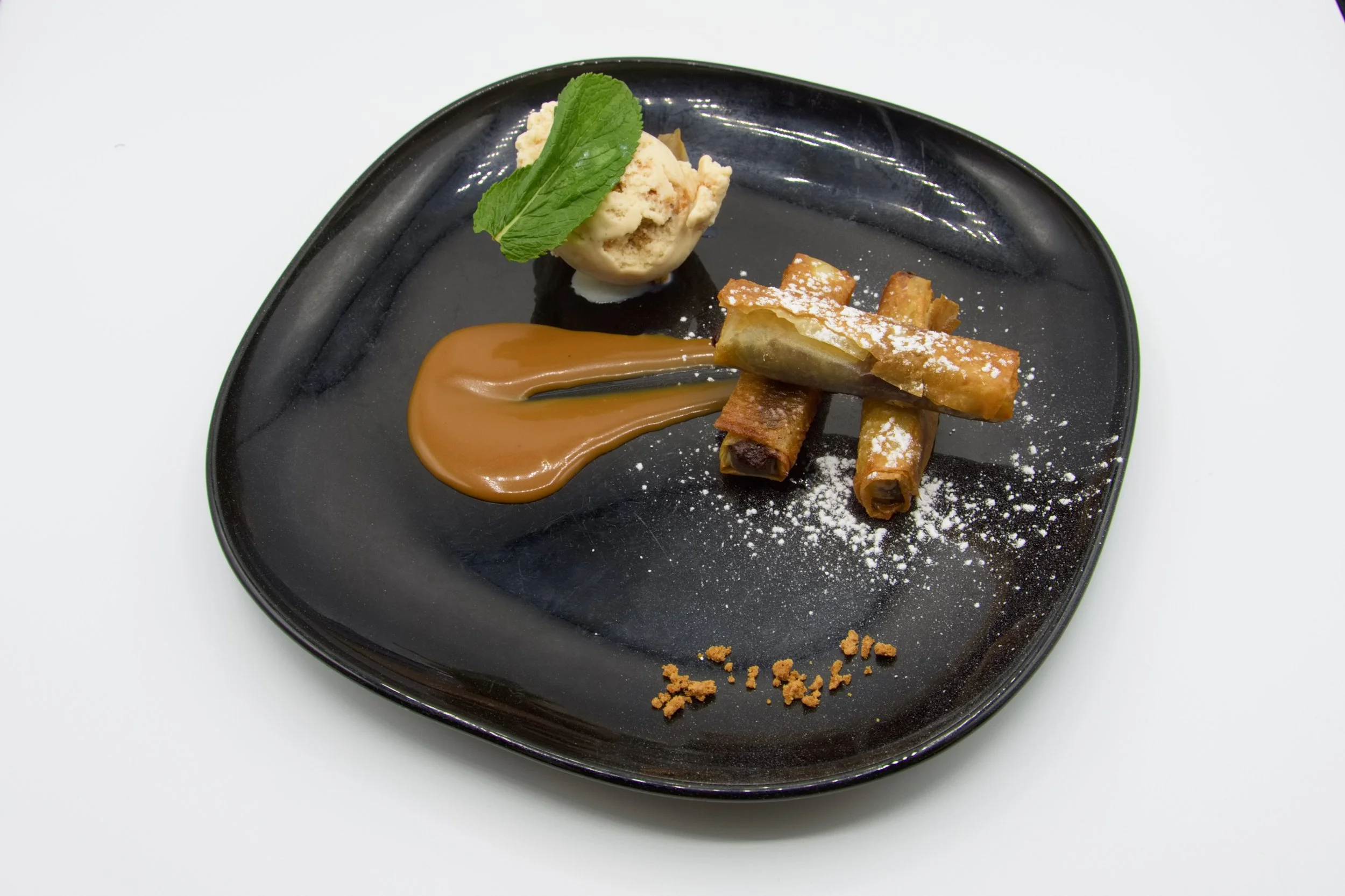Dessert élégant sur une assiette noire avec des rouleaux frits, une boule de glace avec une feuille de menthe, une sauce caramel et une pincée de sucre glace.